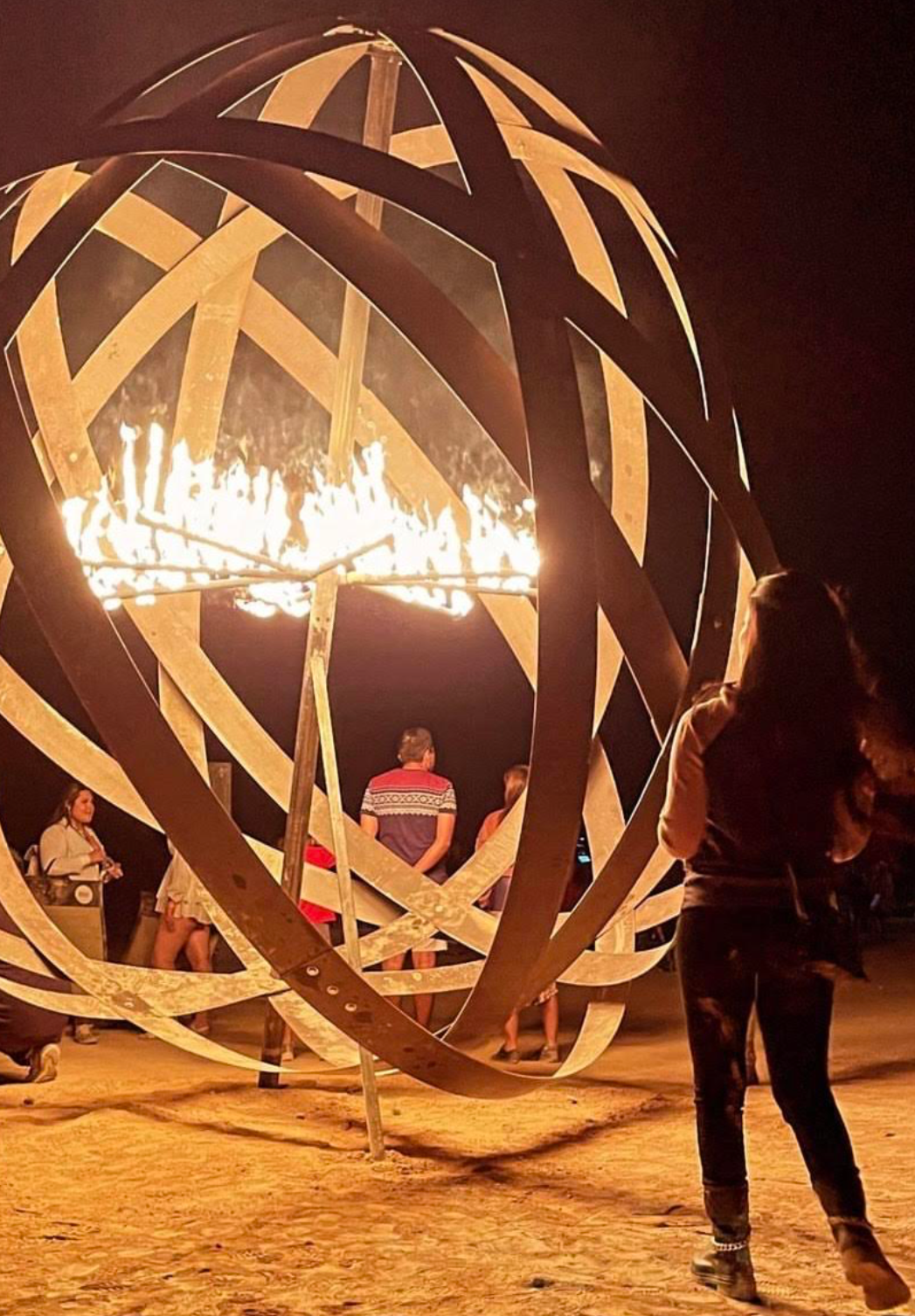 A large outdoor fire sculpture with a spherical, open geometric metal frame. Several people are gathered around, watching the fire burning inside the sculpture during nighttime.