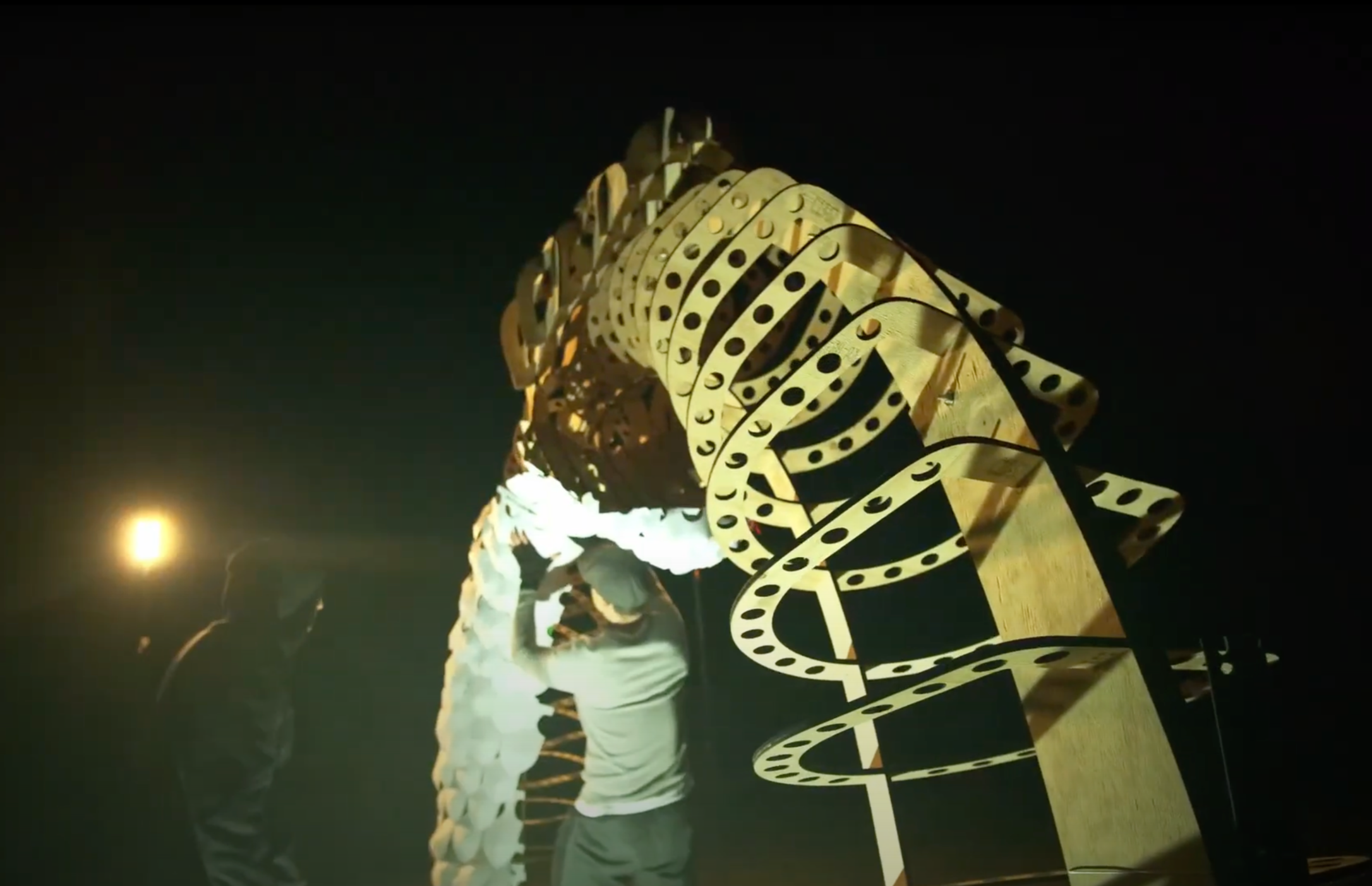 Two people are engaging with a large, illuminated, black-and-yellow sculpture of a snake or dragon at night.