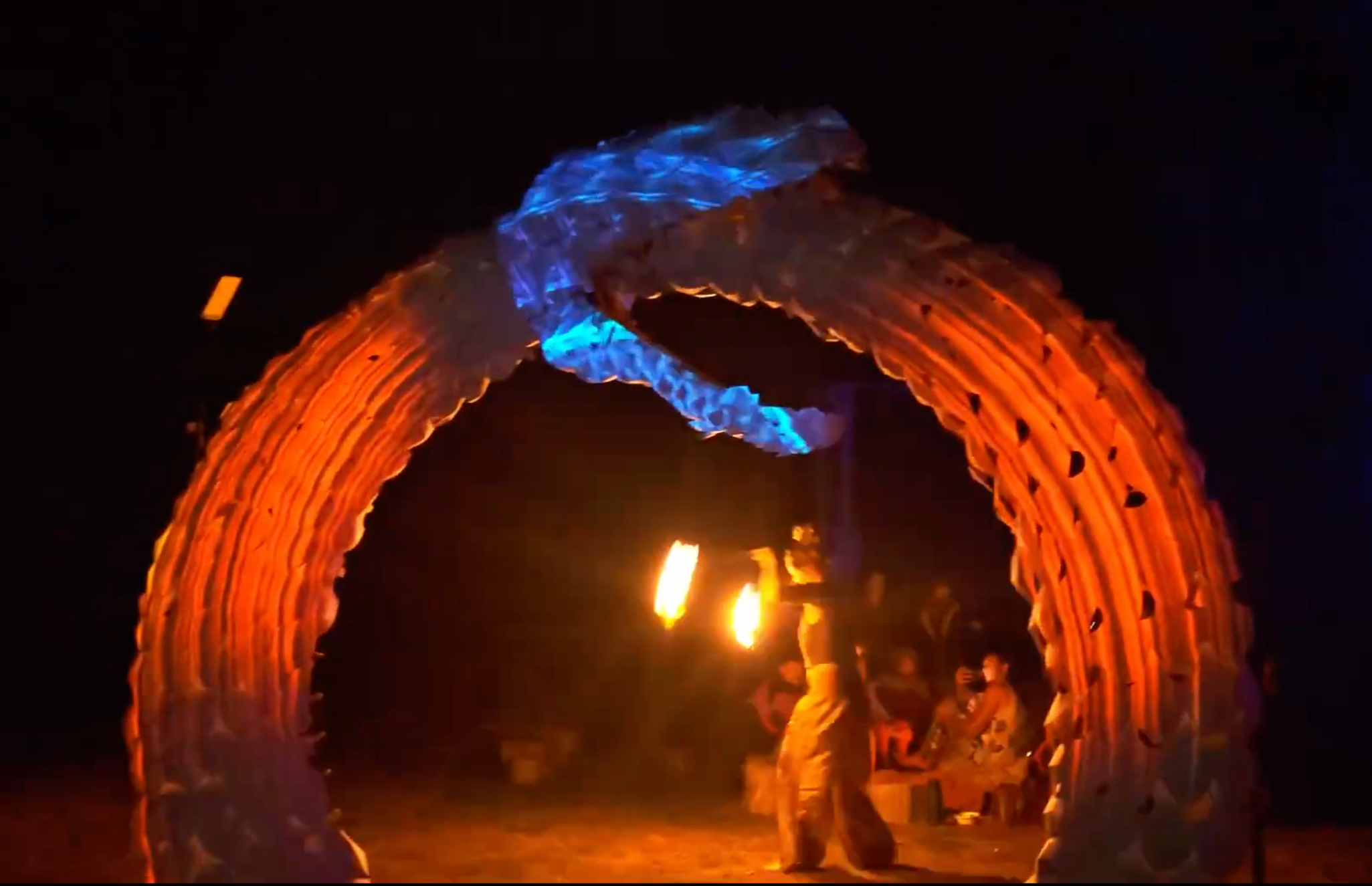People gather around a large bonfire at night, with a metal sculpture of a dragon above it, illuminated by orange and blue lights.