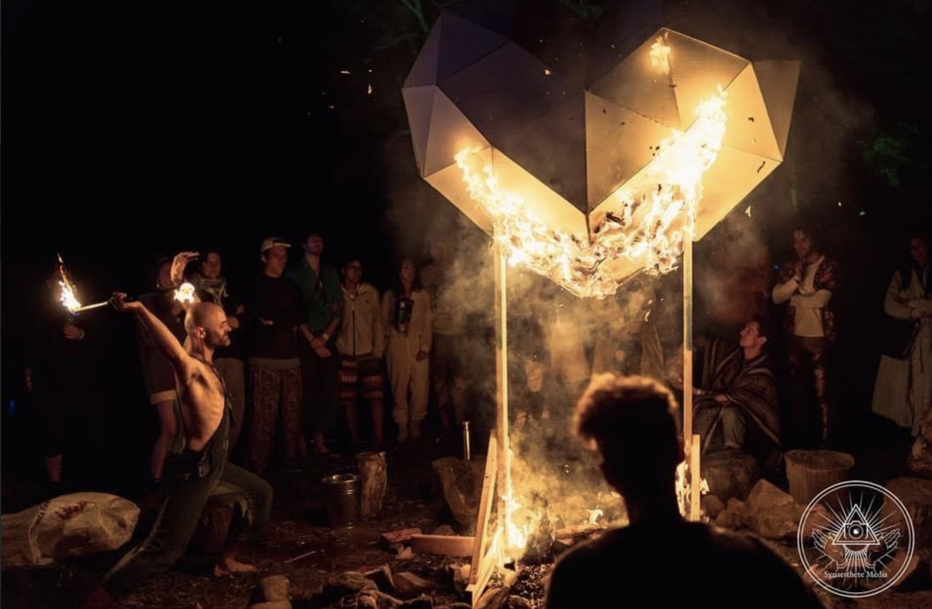 People gathered around a fire in a wooden structure during night, with some individuals observing and one person actively tending the flames.