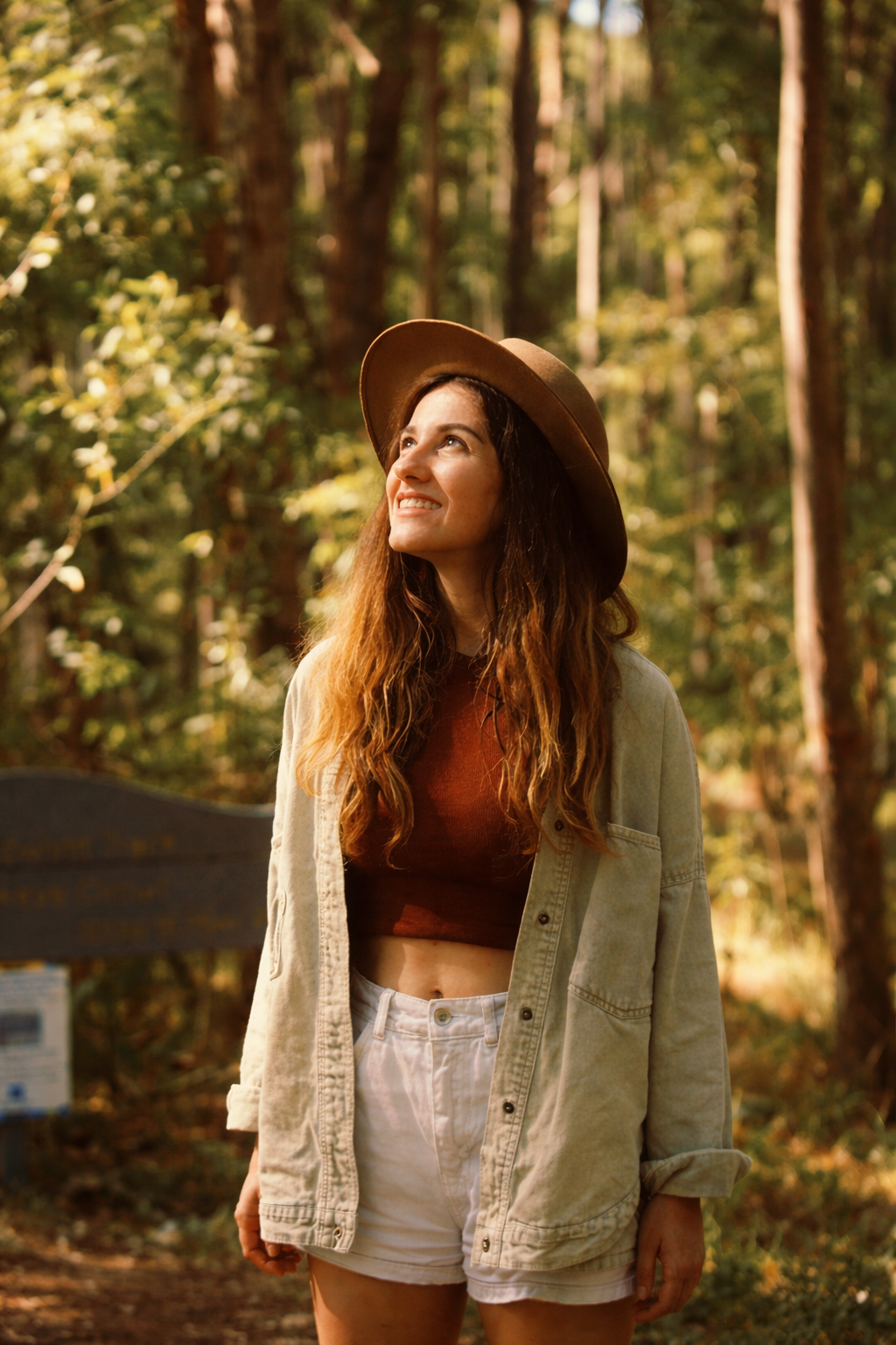 A woman with long, wavy hair wearing a brown hat, a cropped maroon top, a light-colored jacket, and white shorts, standing in a forest and looking upward with a smile.