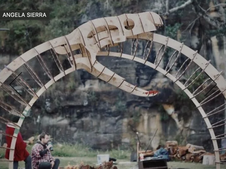 Large wooden dragon sculpture outdoors with two people in the foreground, one sitting and one standing, near a river or stream.