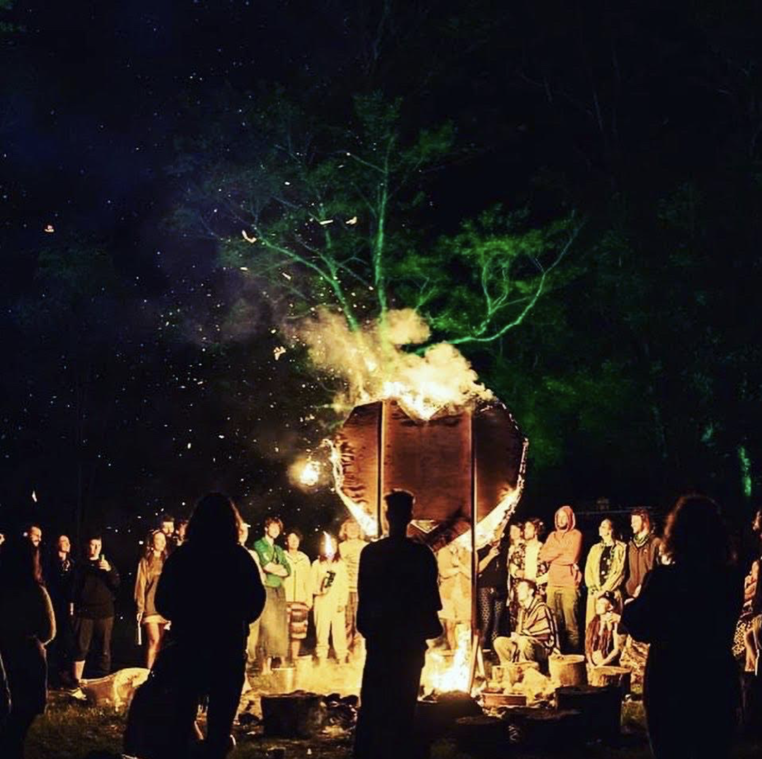 People gathered around a large burning paper lantern at night, with green fireworks illuminating the sky above.