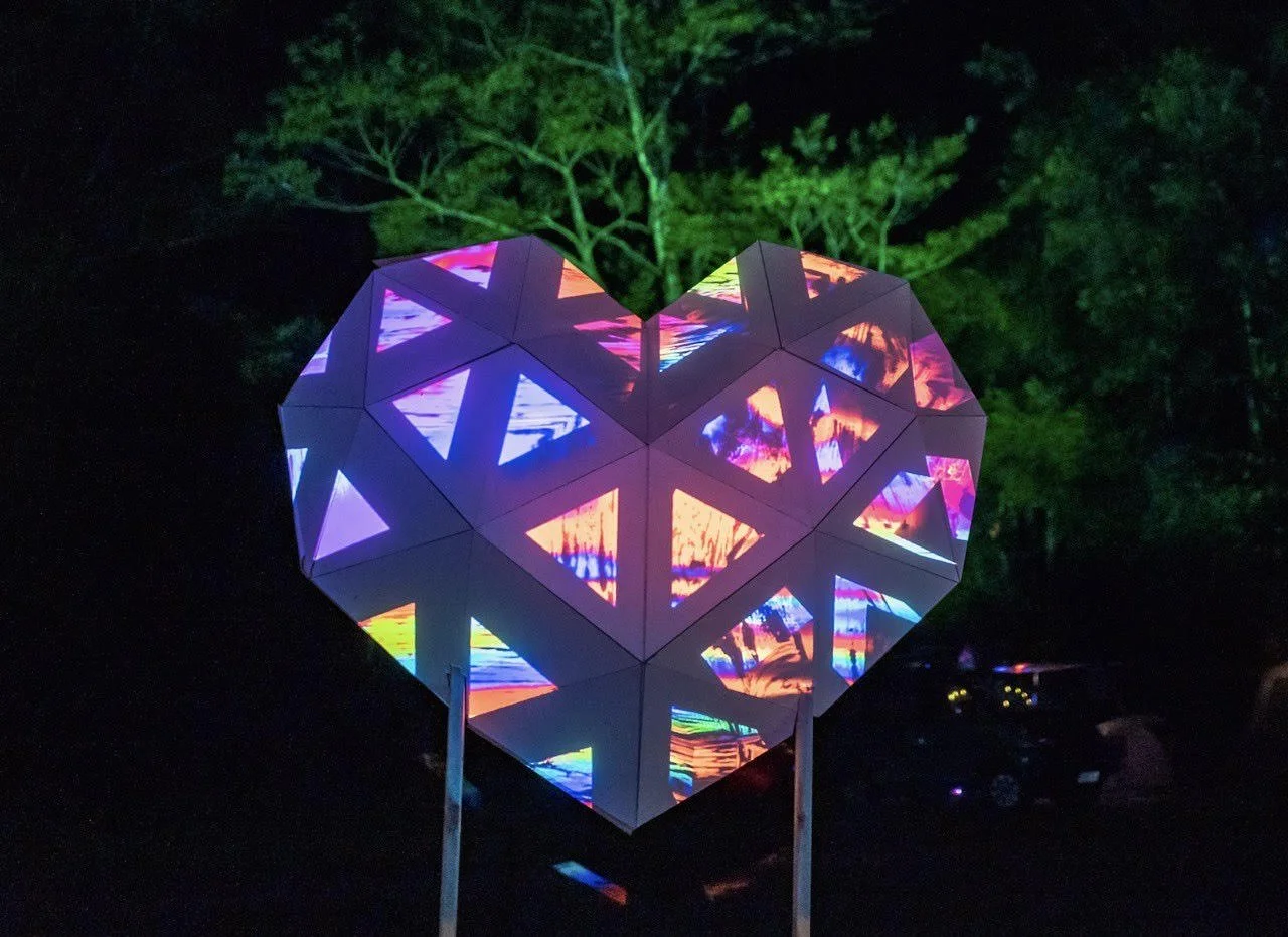 A glowing, heart-shaped structure made of triangular panels with colorful, abstract patterns. It is outdoors at night with trees in the background.
