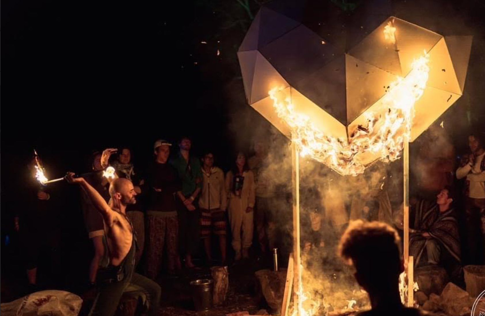 A group of people gathered around a large wooden structure that is on fire, possibly during a festival or ritual. The scene is set at night with the flames illuminating the surroundings.