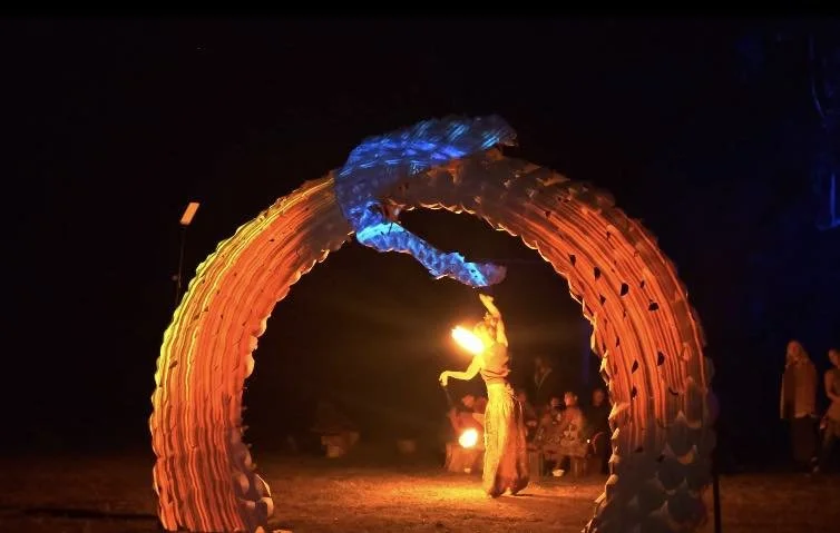 A person performing a fire dance under a large, colorful, illuminated arch at night.