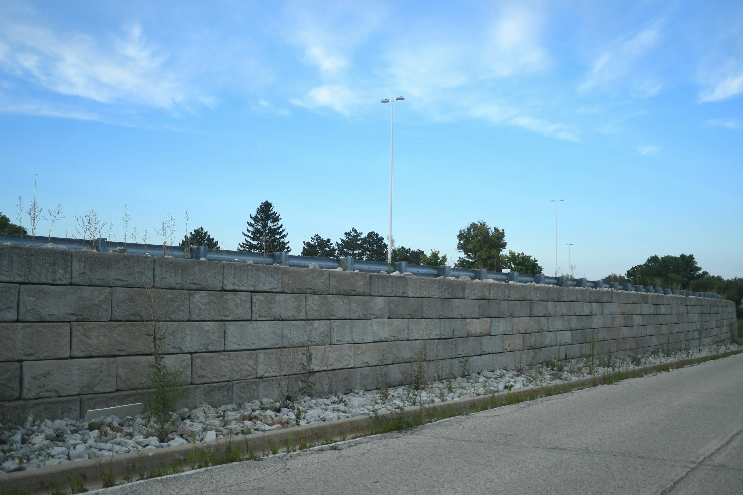 View of a stone retaining wall beside a road, with young trees and shrubs planted behind it, under a blue sky with wispy clouds.