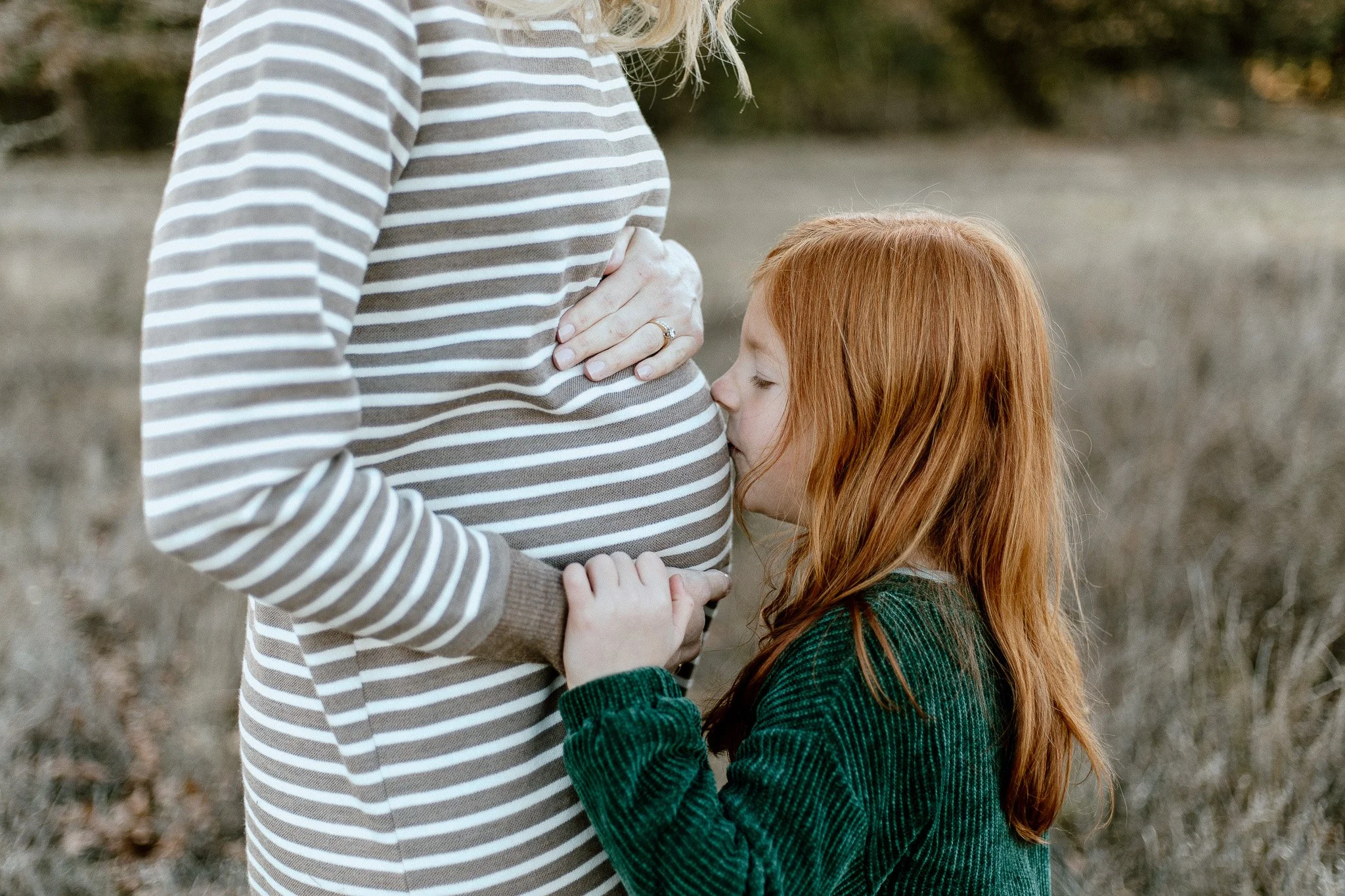 A young girl with red hair embracing and kissing her mother's pregnant stomach outdoors. The woman is wearing a striped long-sleeve shirt and has her hand resting on her stomach. The background is blurred with natural outdoor scenery.