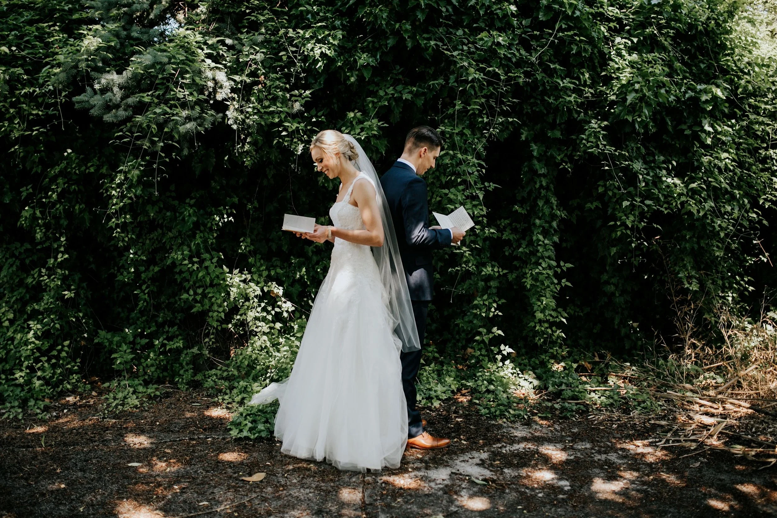 A bride and groom standing back to back outdoors among green foliage, reading from books, with the bride smiling and wearing a white wedding dress and veil, and the groom in a dark suit.