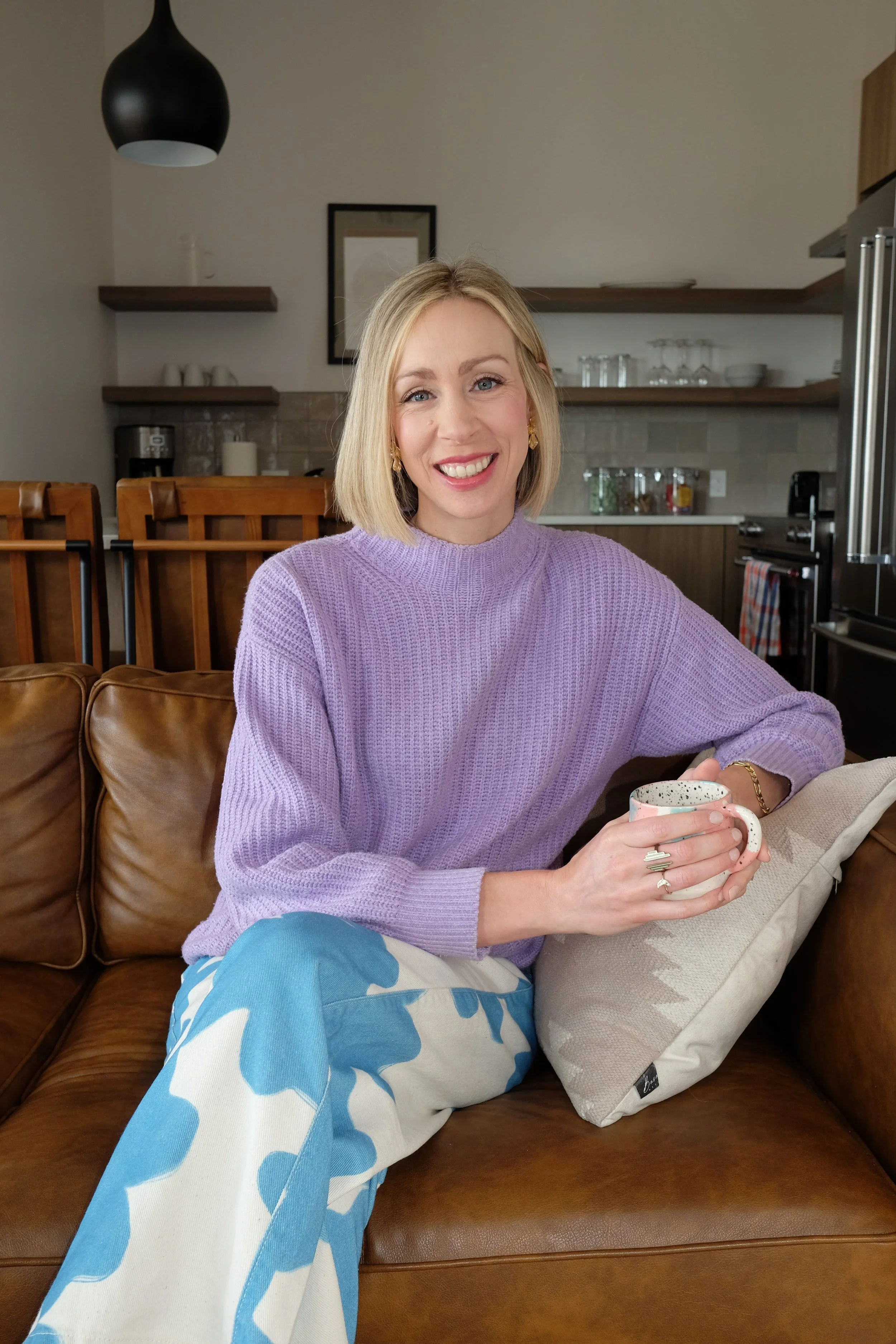 A woman with blonde hair wearing a lavender sweater, sitting on a leather couch in a kitchen, smiling and holding a mug in her hands.