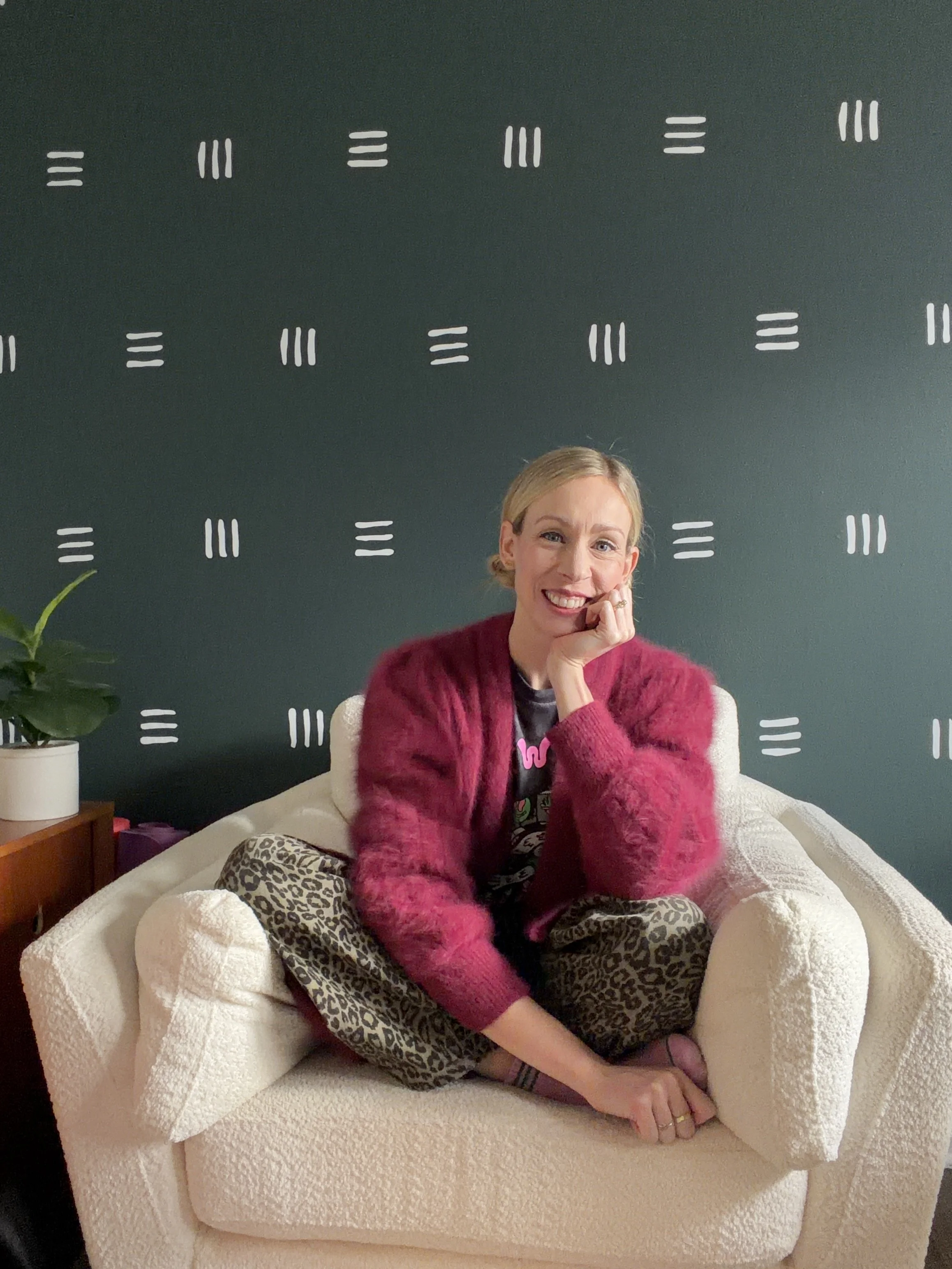 A woman sitting on a beige armchair with leopard print pants, wearing a fuzzy pink sweater, smiling and resting her chin on her hand in a room with a dark green wall and white patterned decorations.