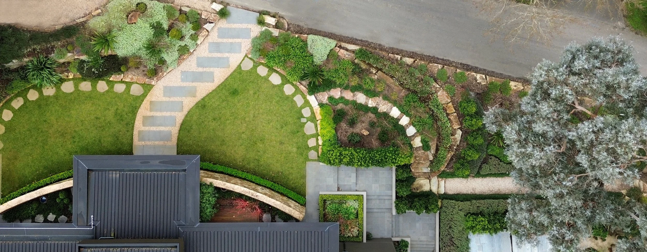Aerial view of a landscaped front yard with a curved stone pathway, green lawn, various bushes, and trees next to a driveway.
