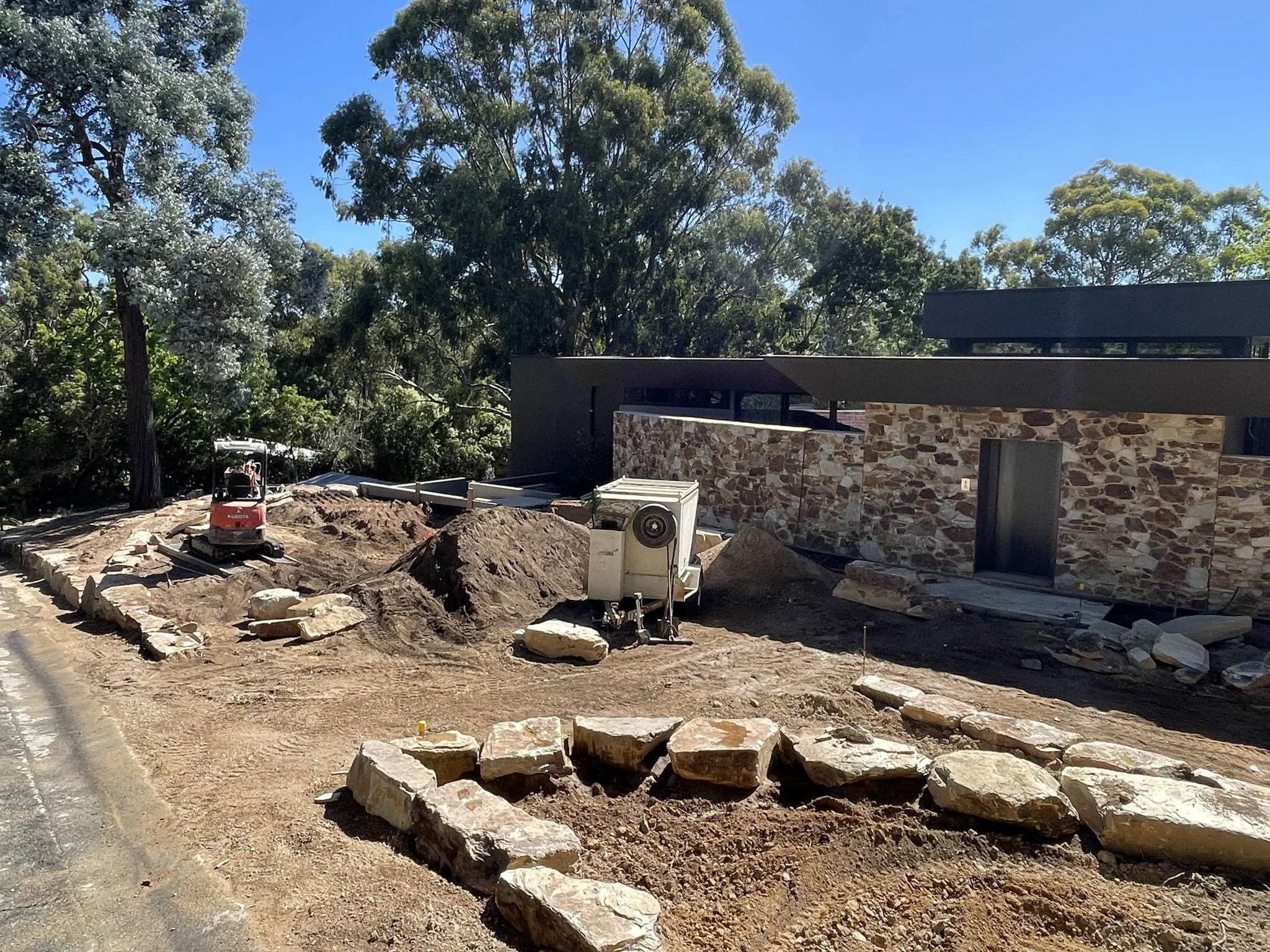A construction site with large rocks and dirt, partially built modern house with stone exterior, trees in the background, and bright blue sky.
