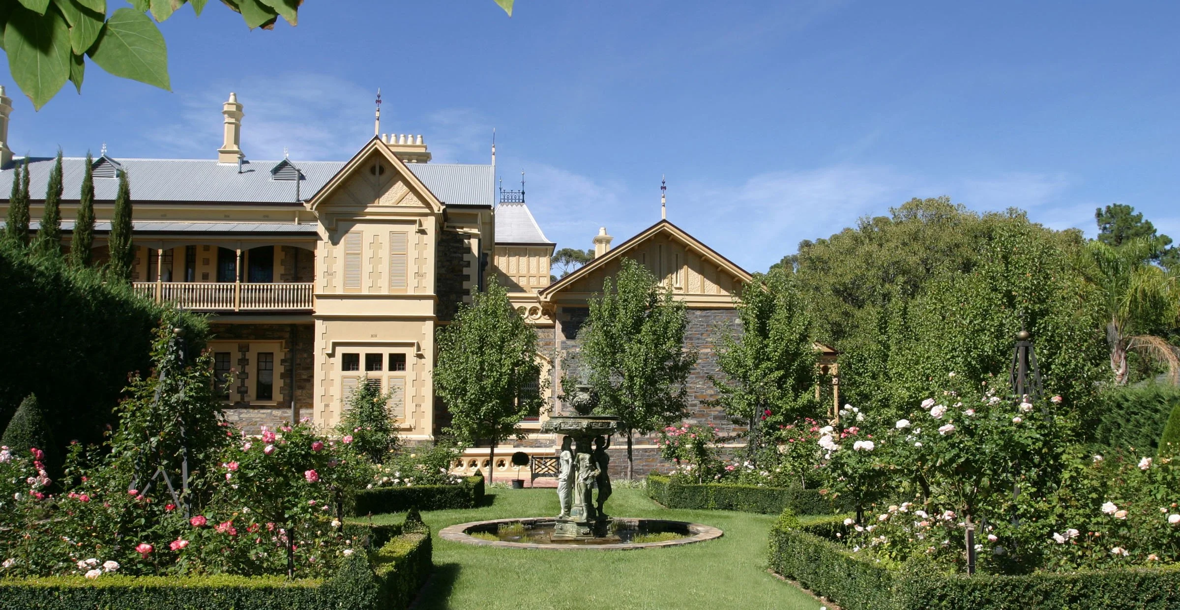 A large, historic house with a ornate facade, surrounded by a manicured garden with a fountain and blooming roses. The house has multiple gables with decorative trim and tall chimneys, set against a bright blue sky.