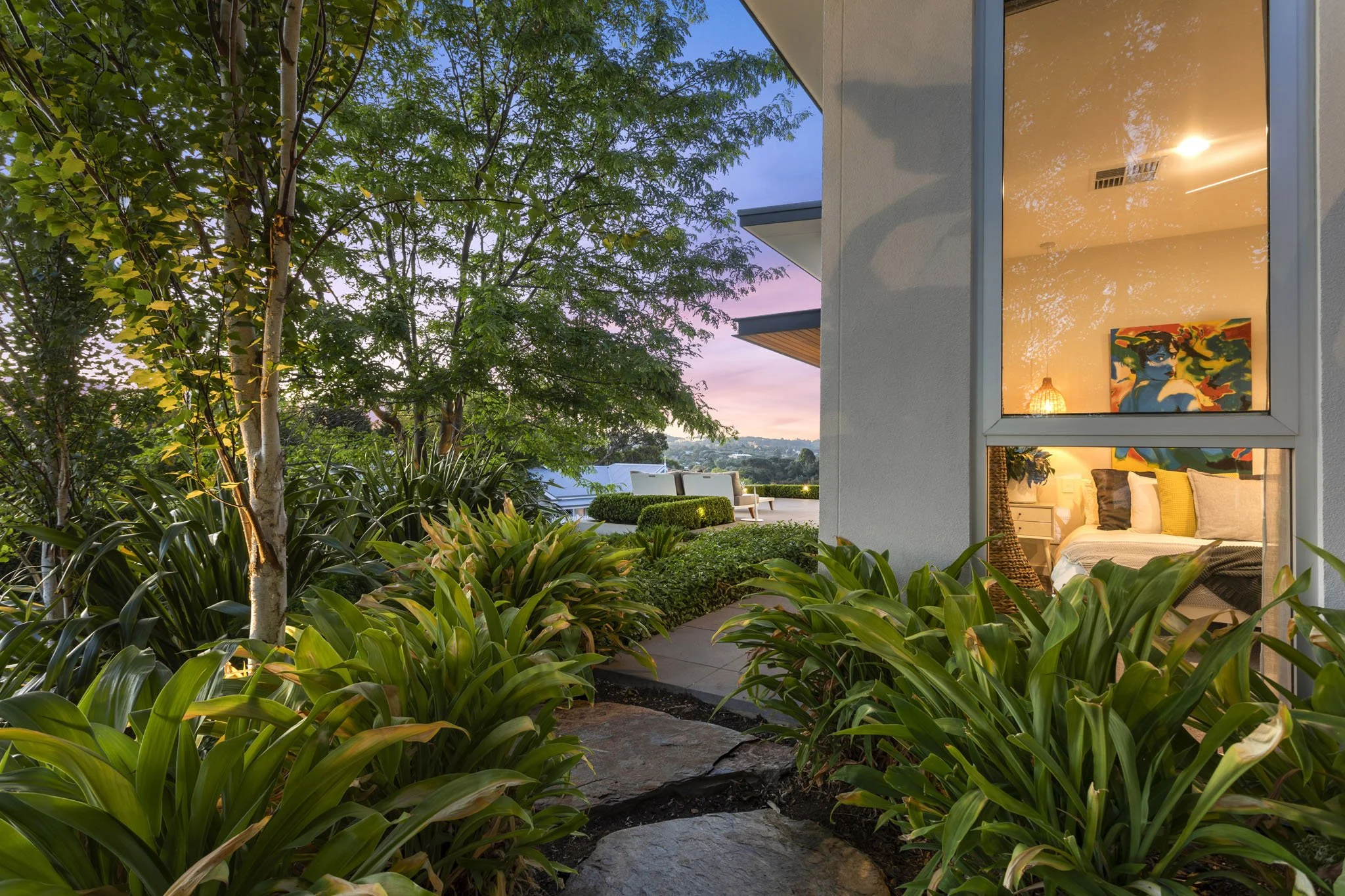 Exterior view of a modern house with large glass windows illuminated from inside, surrounded by lush green plants and trees during dusk.