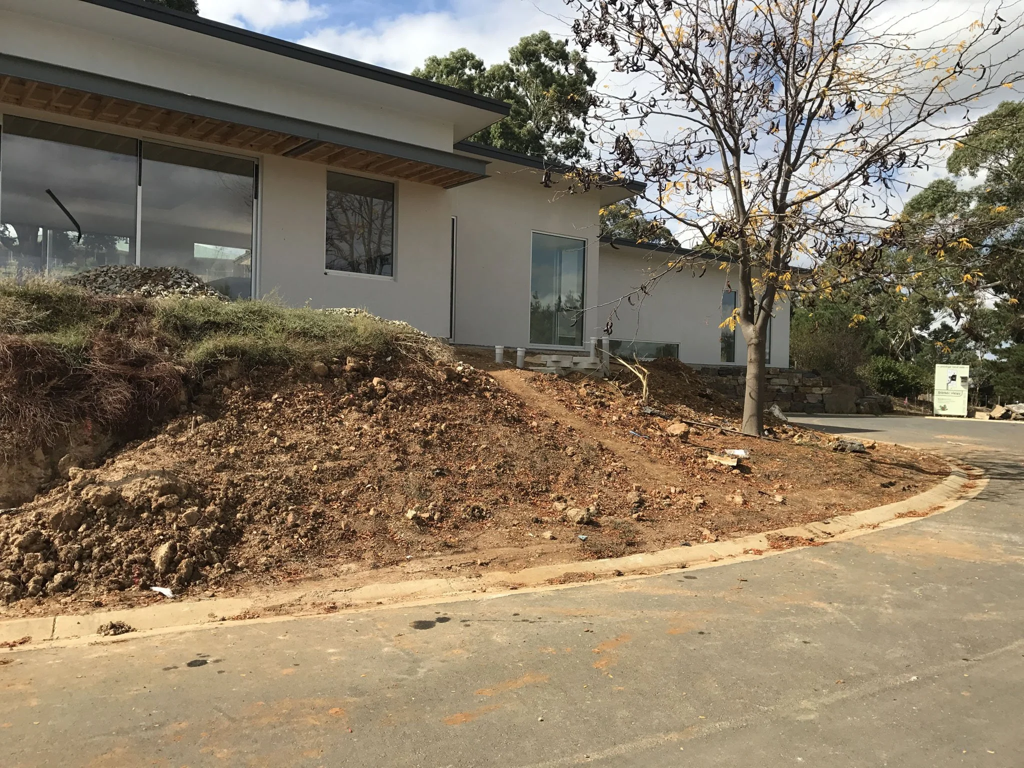 Modern single-story house with large windows, surrounded by a dirt landscape, trees, and a partly cloudy sky.