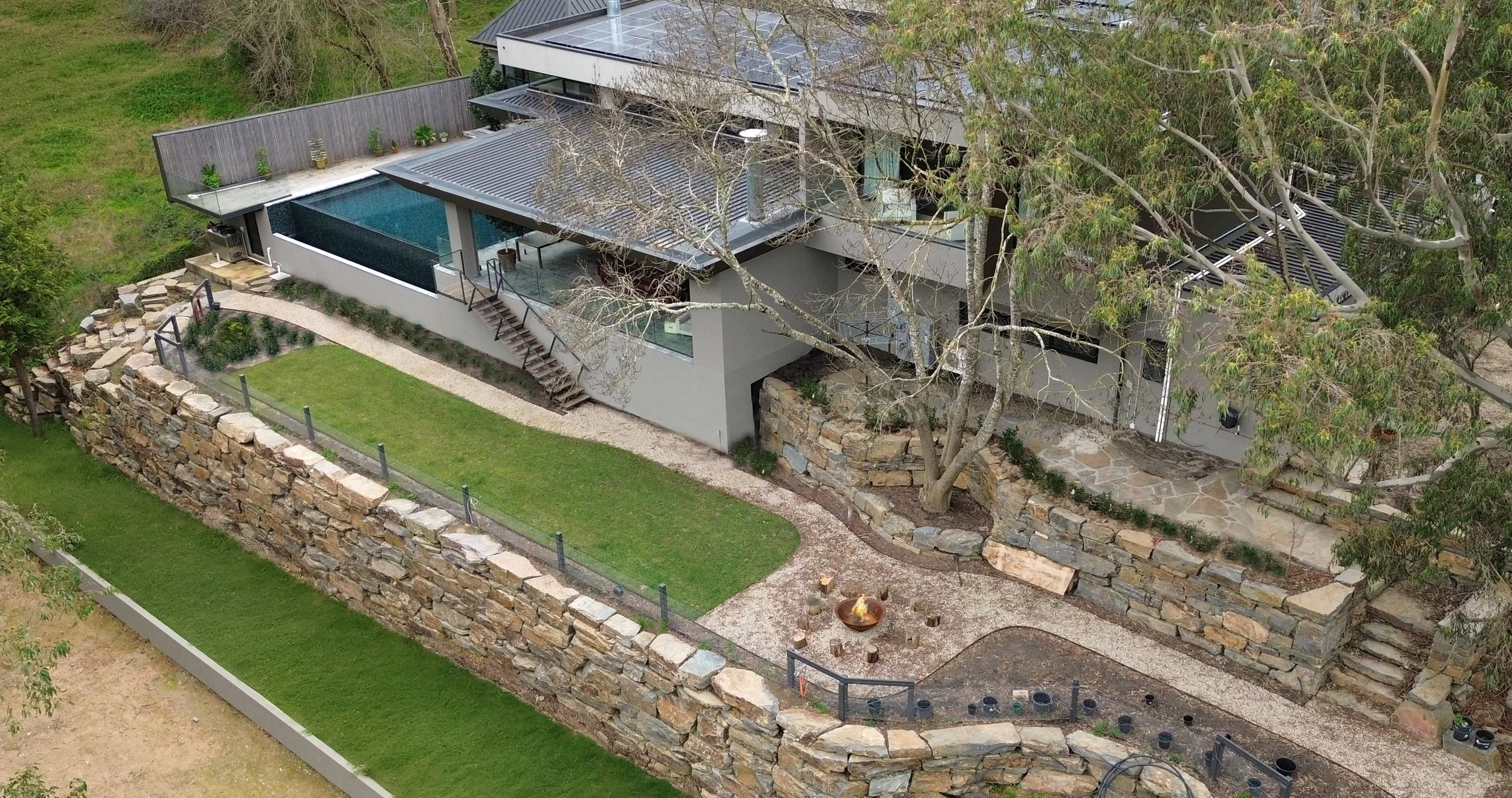 A modern house with multiple levels and large glass windows, featuring a swimming pool and outdoor seating areas, surrounded by a stone wall and greenery.