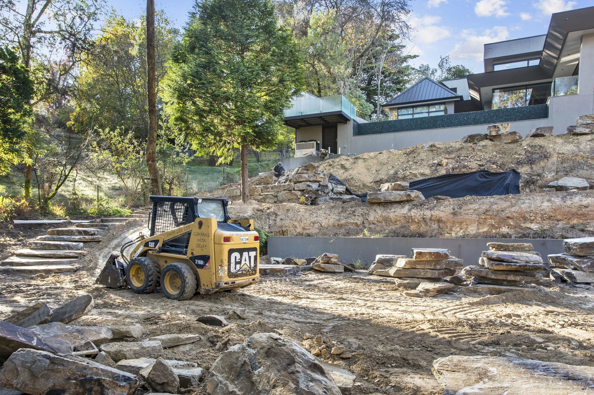 Construction site with dirt and rocks, a small yellow CAT backhoe, and workers in the background. Modern house with large windows and a glass fence on a hillside surrounded by trees.