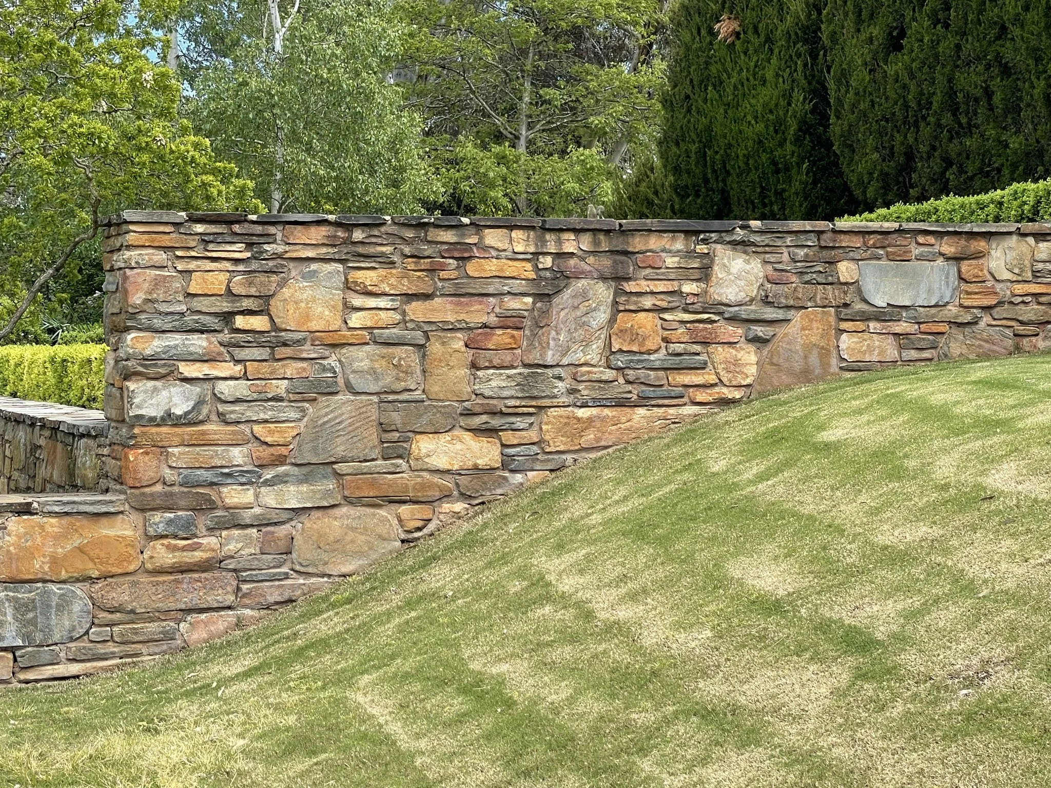 A stone wall with a textured surface, bordered by a green lawn, and trees with green foliage in the background.