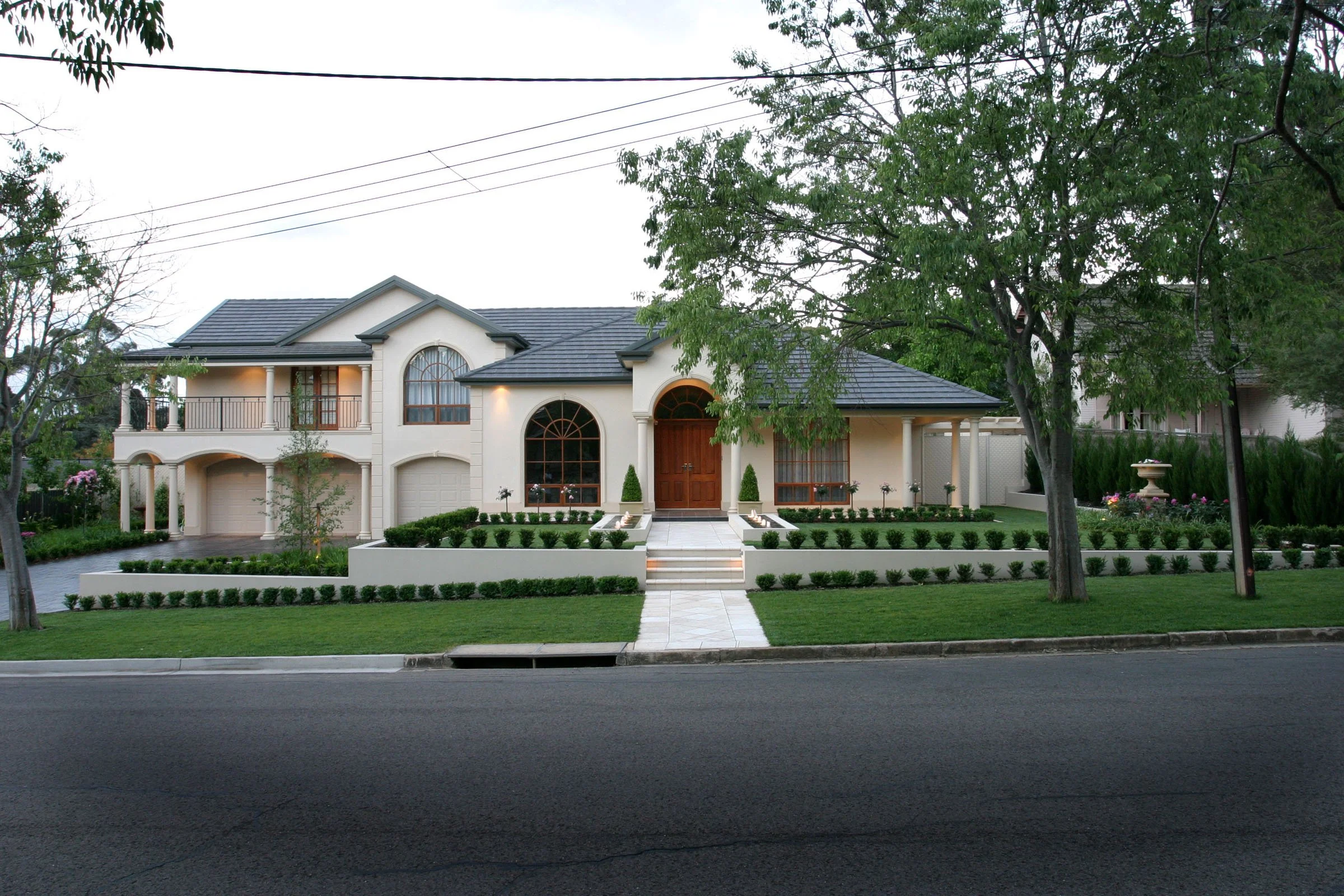 Front view of a large, elegant house with a well-manicured lawn, lined with small bushes, and a walkway leading to the front door. There are tall trees on both sides of the house, and a sidewalk and street in the foreground.