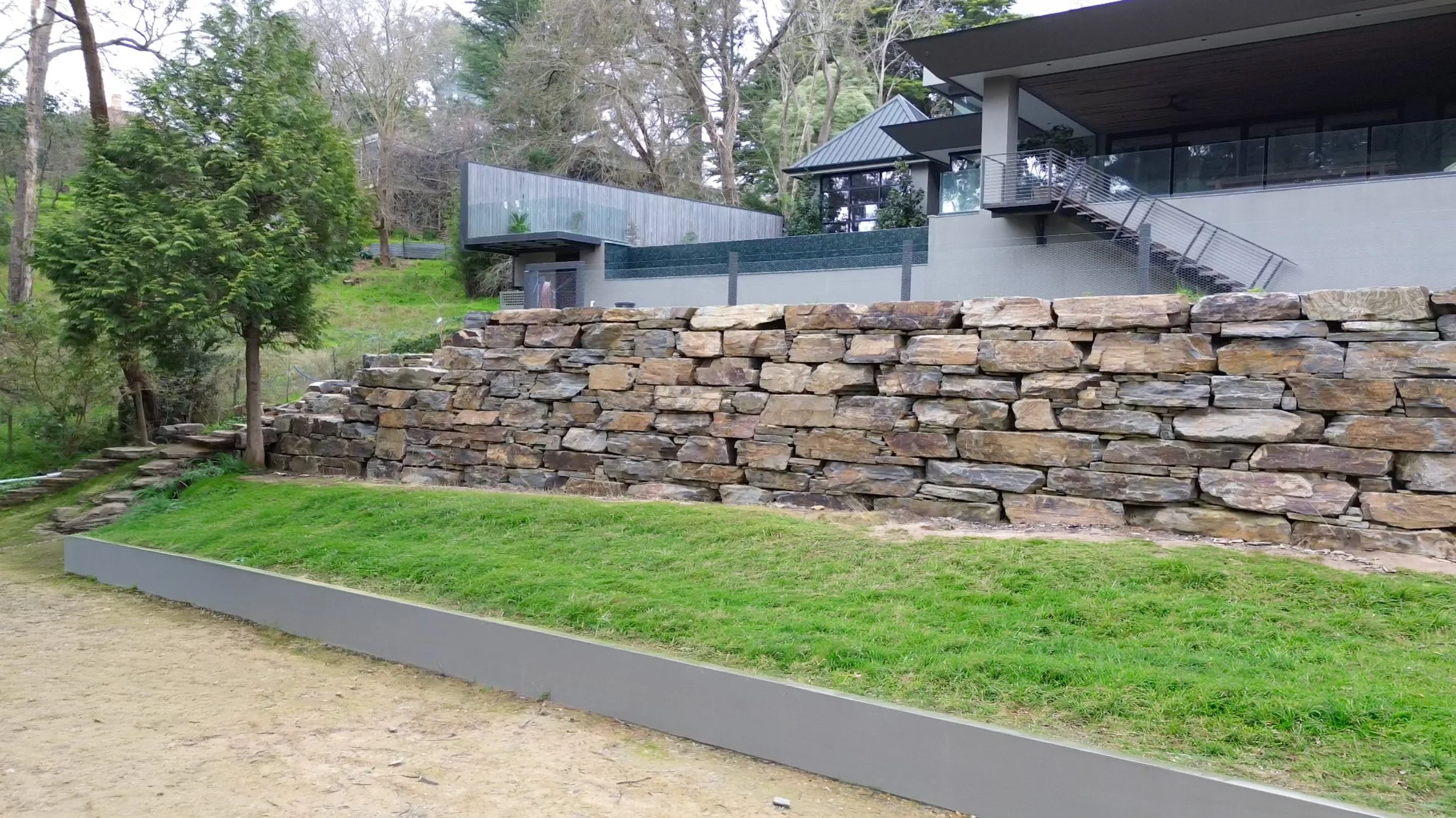 Modern house built on a hillside with black metal stairs, glass railings, and a stone retaining wall, surrounded by trees and green lawn.
