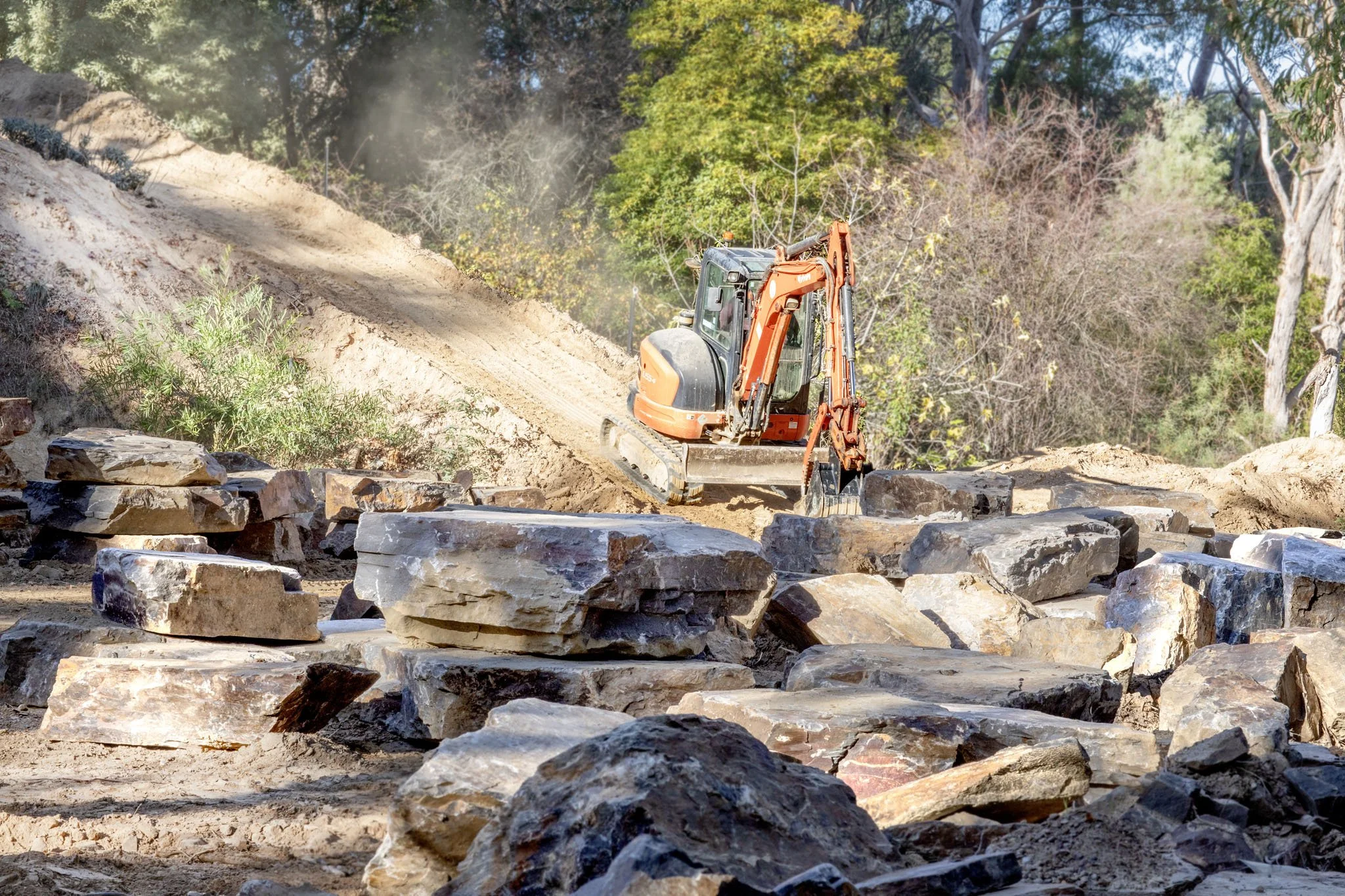Construction site with a small excavator working on a dirt hillside surrounded by trees, with rocks in the foreground and dust in the air.
