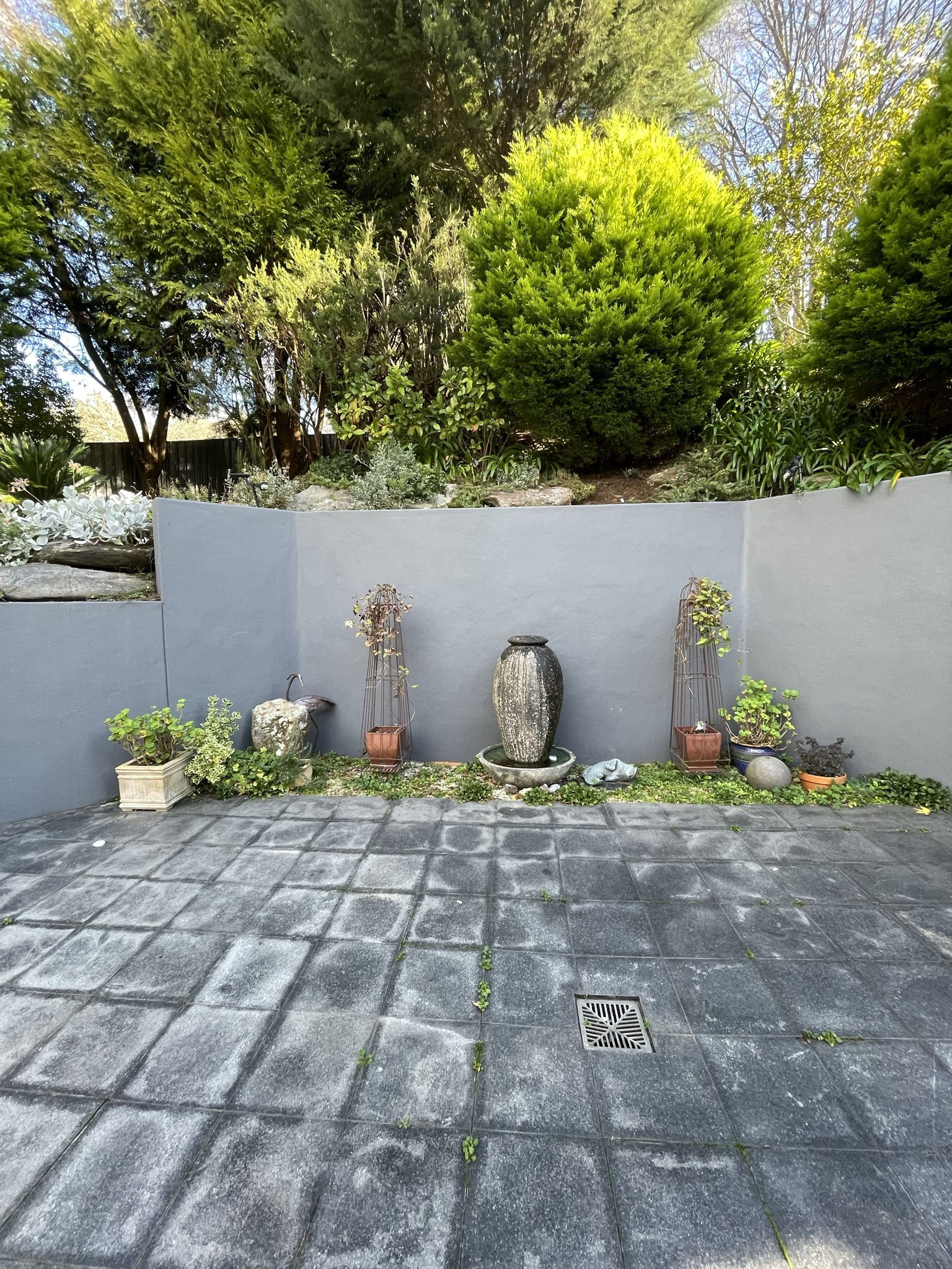 A backyard with a gray concrete patio featuring visible grout lines and a drainage grate. In front of a blue wall, there are potted plants, decorative stones, and a tall stone garden fountain. Behind the wall, there is a garden bed with various shrubs, plants, and rocks, and a large green bush. In the background, there are tall trees with mostly bare branches and some greenery, under a partly cloudy sky.