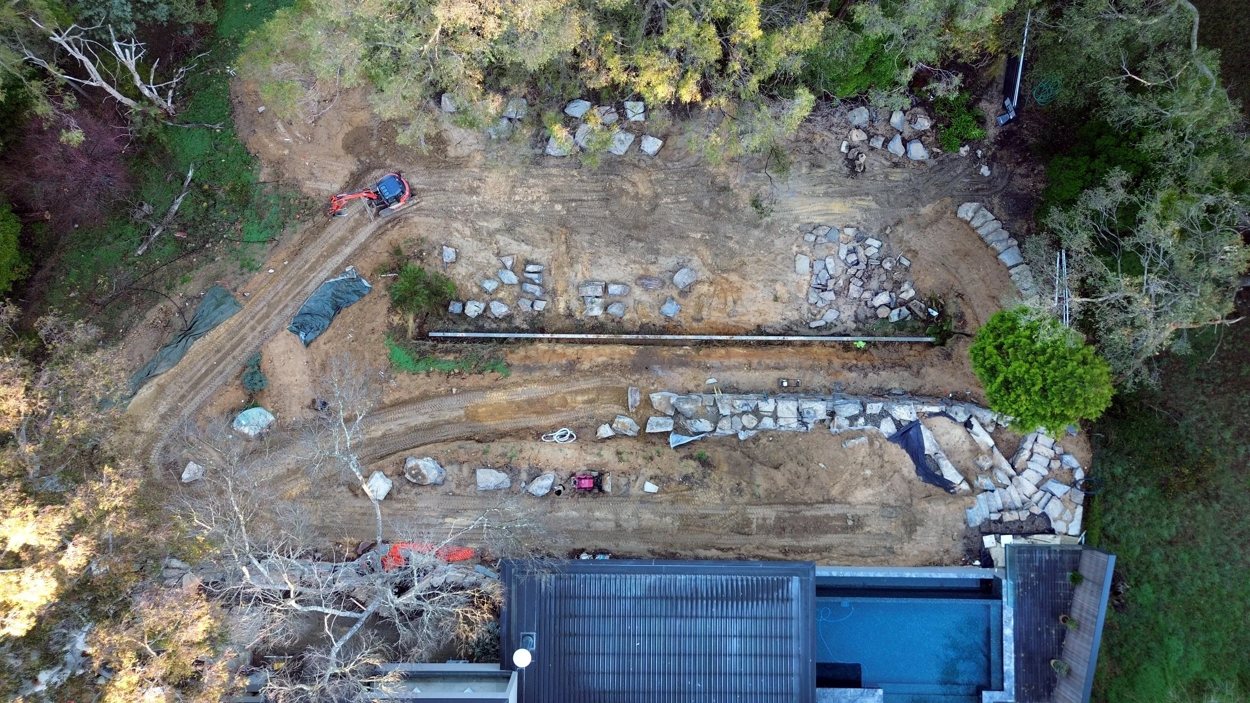 Aerial view of a construction site with a dirt road, large rocks, construction equipment, and surrounding trees.