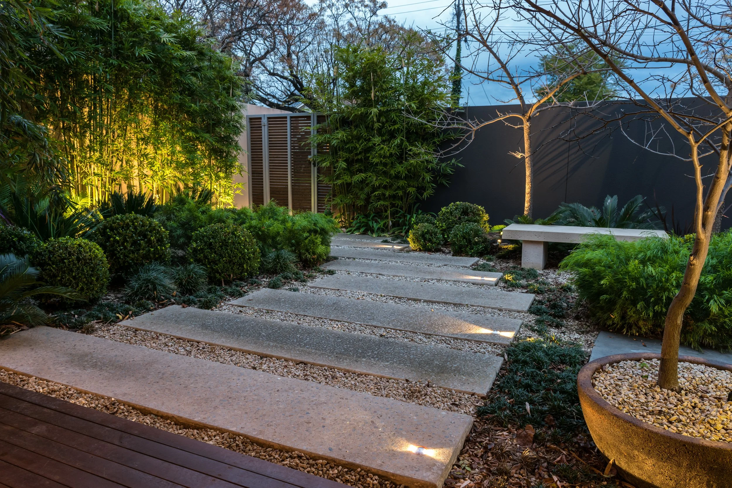 A well-lit modern backyard garden during dusk, featuring a stone pathway, lush green shrubs, trees, and a black fence, with some trees bare of leaves and outdoor ambient lighting.