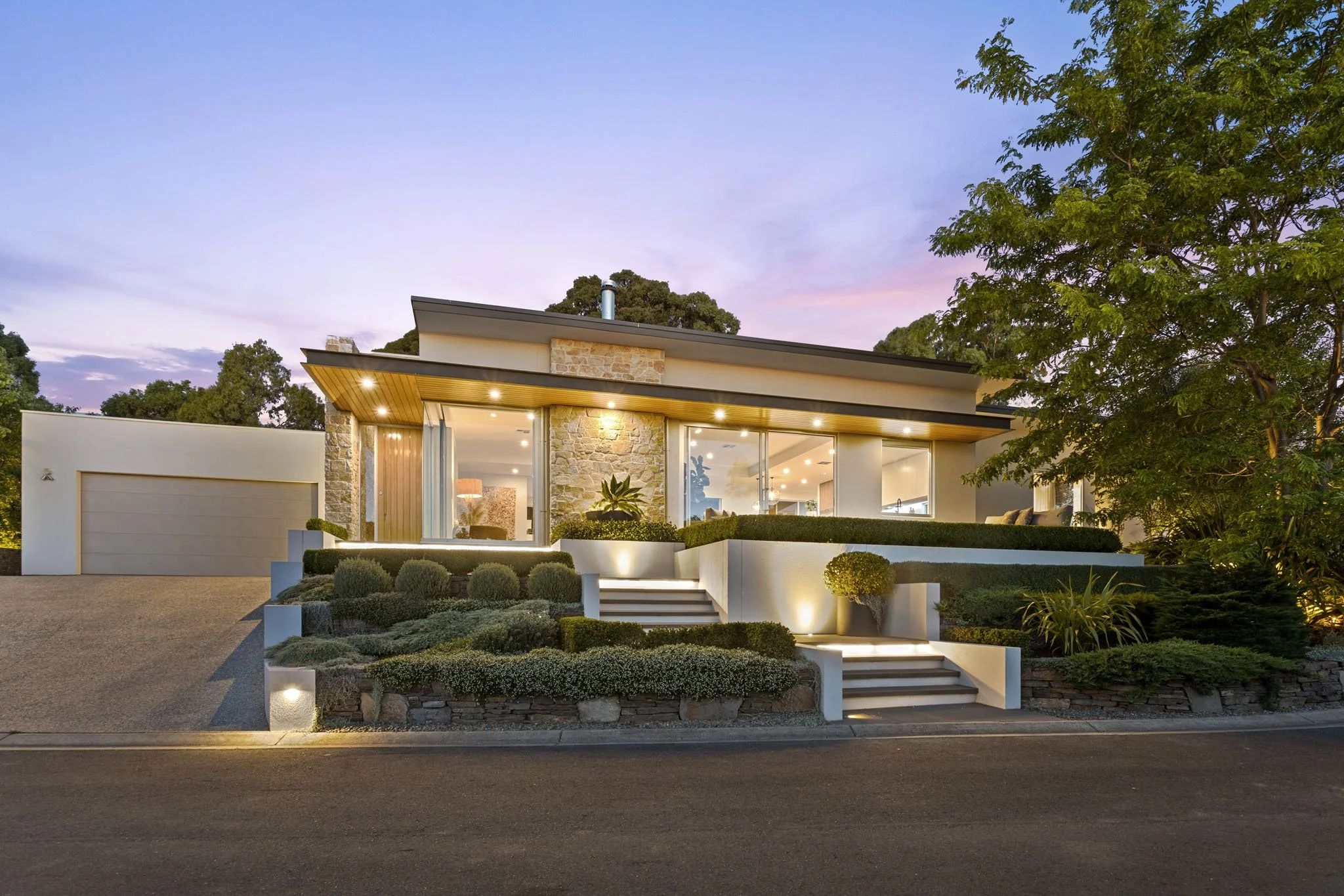 Modern house with large glass windows, stone accents, and exterior lighting at dusk, surrounded by landscaped garden and trees.