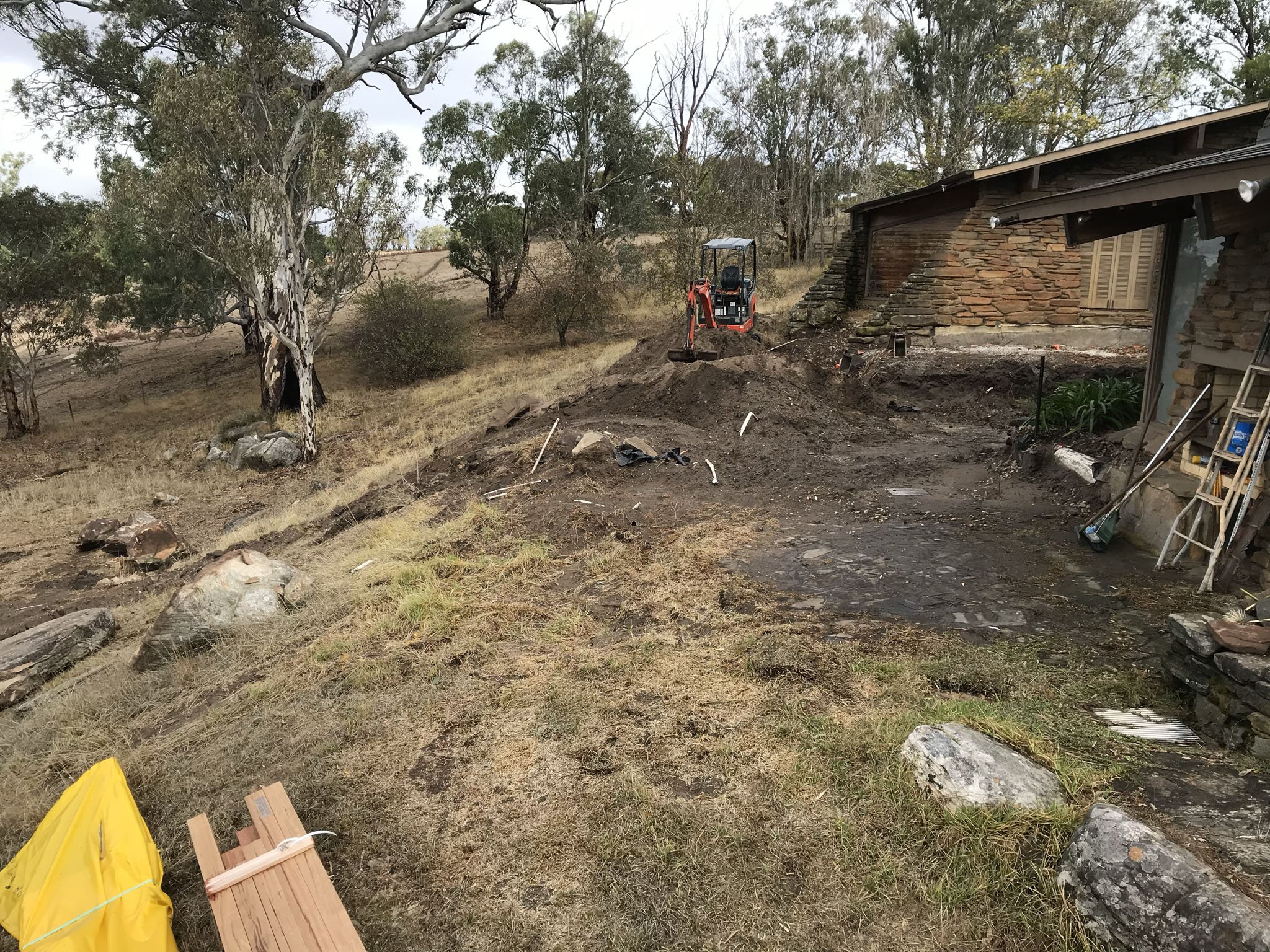 Construction site with an excavator digging near a house, surrounded by trees and construction materials on the ground.