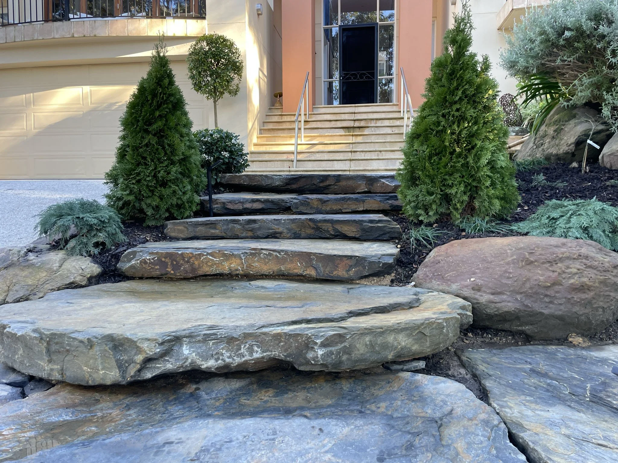 Exterior of a house with a staircase leading to a front door, surrounded by green shrubs and trees, with rocks lining the path.