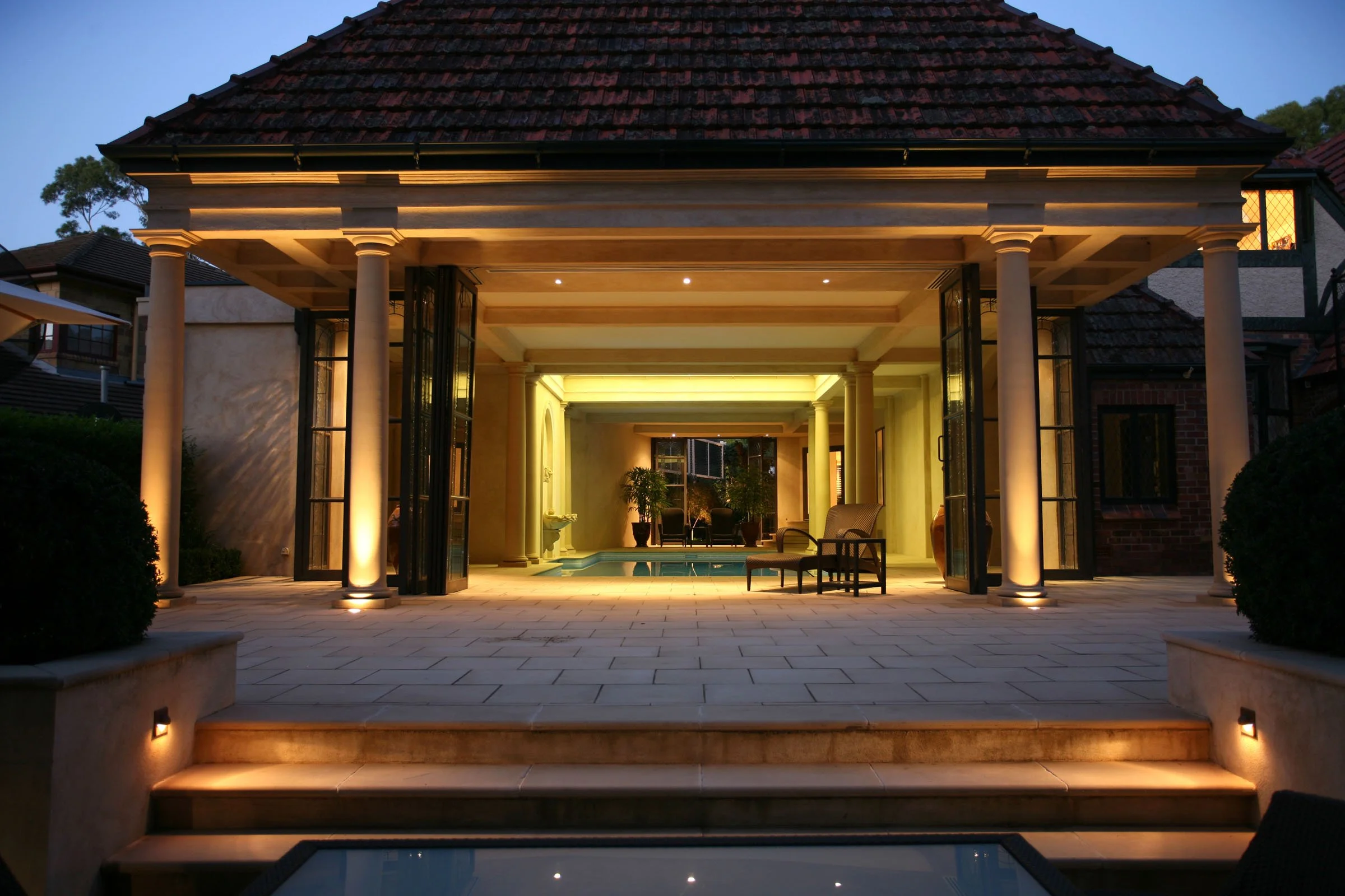 Backyard patio with a covered seating area, outdoor pool, and stairs leading to the patio, illuminated with warm lighting at dusk.
