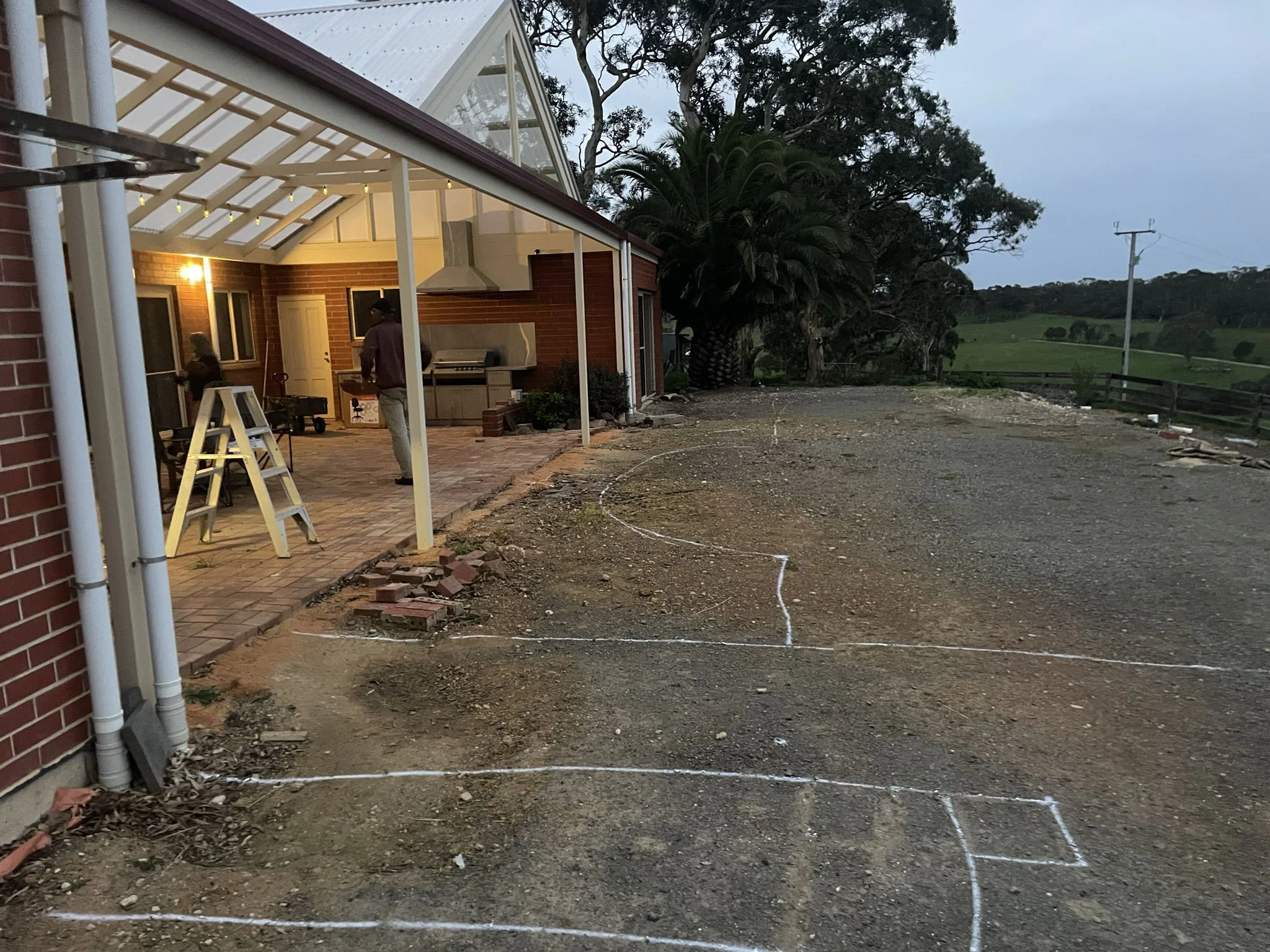 A house with a brick exterior and a porch area under construction, with a ladder and construction tools visible. The yard is disturbed with dirt and markings on the ground, and there are trees and a power line in the background.