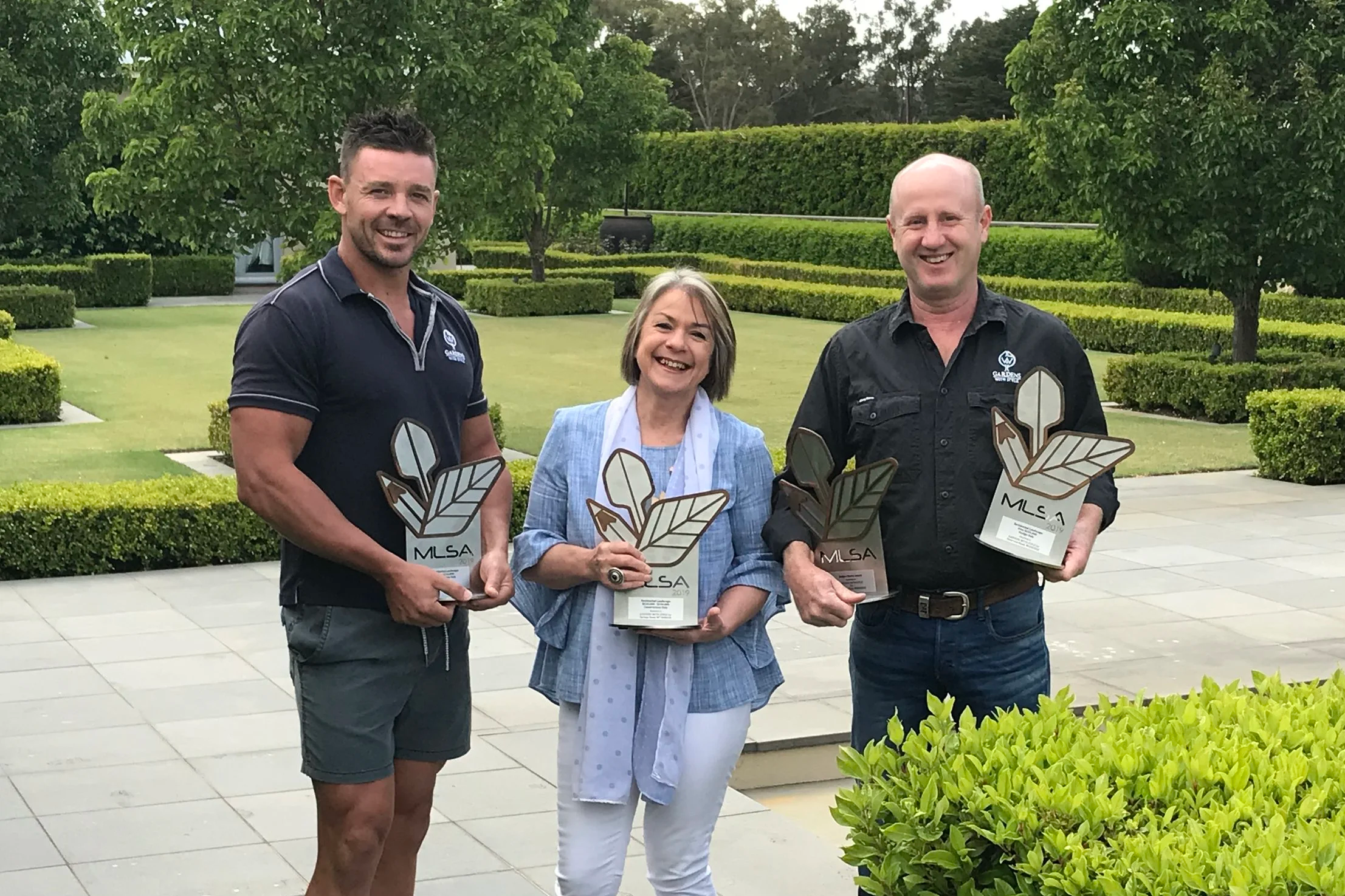 Three people standing outdoors holding awards with an ornamental garden in the background, smiling at the camera.