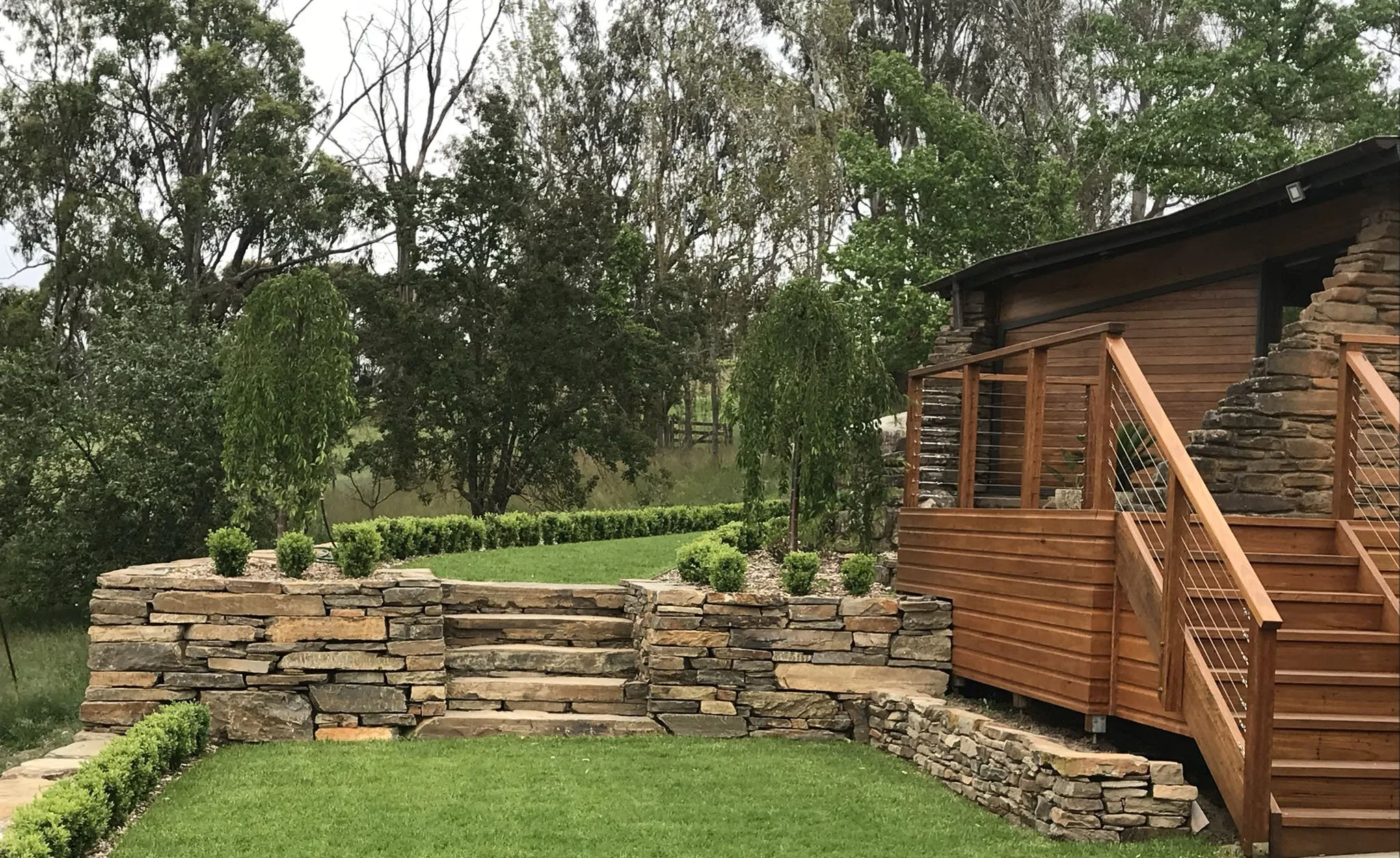 A backyard with a stone retaining wall, neatly trimmed shrubs, green grass, a wooden deck with stairs, and tall trees in the background.