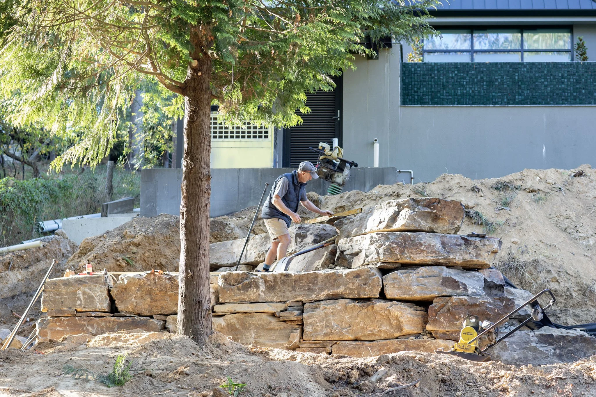 A person working on a construction site outdoors, laying large stone slabs, with a tree in the foreground and a modern building in the background.
