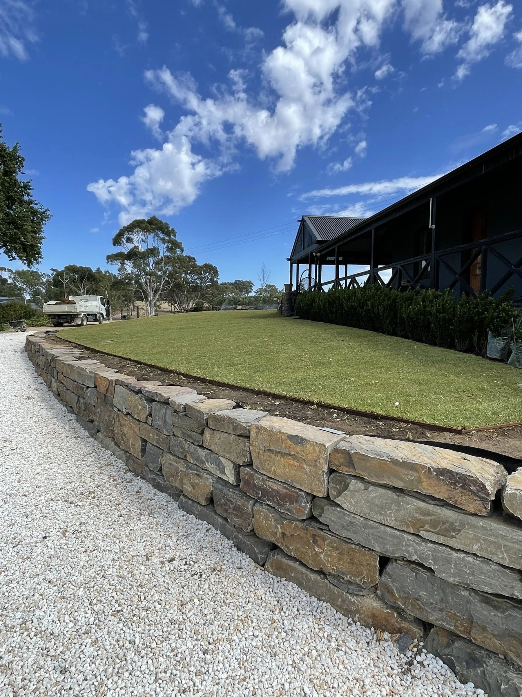 A landscaped yard with a stone retaining wall, green grass, gravel pathway, and a house with a porch and a metal roof under a partly cloudy sky.