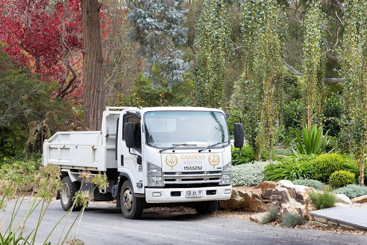 White Isuzu flatbed truck parked on a gravel driveway in a lush garden with trees, bushes, and rocks