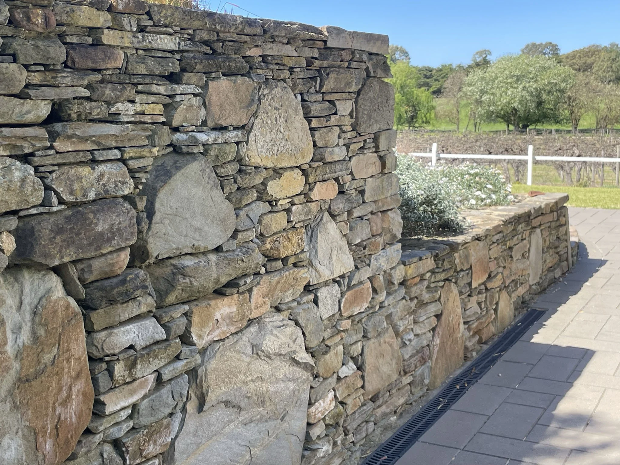 Stone wall with various-sized rocks and a garden with trees and a white fence in the background.