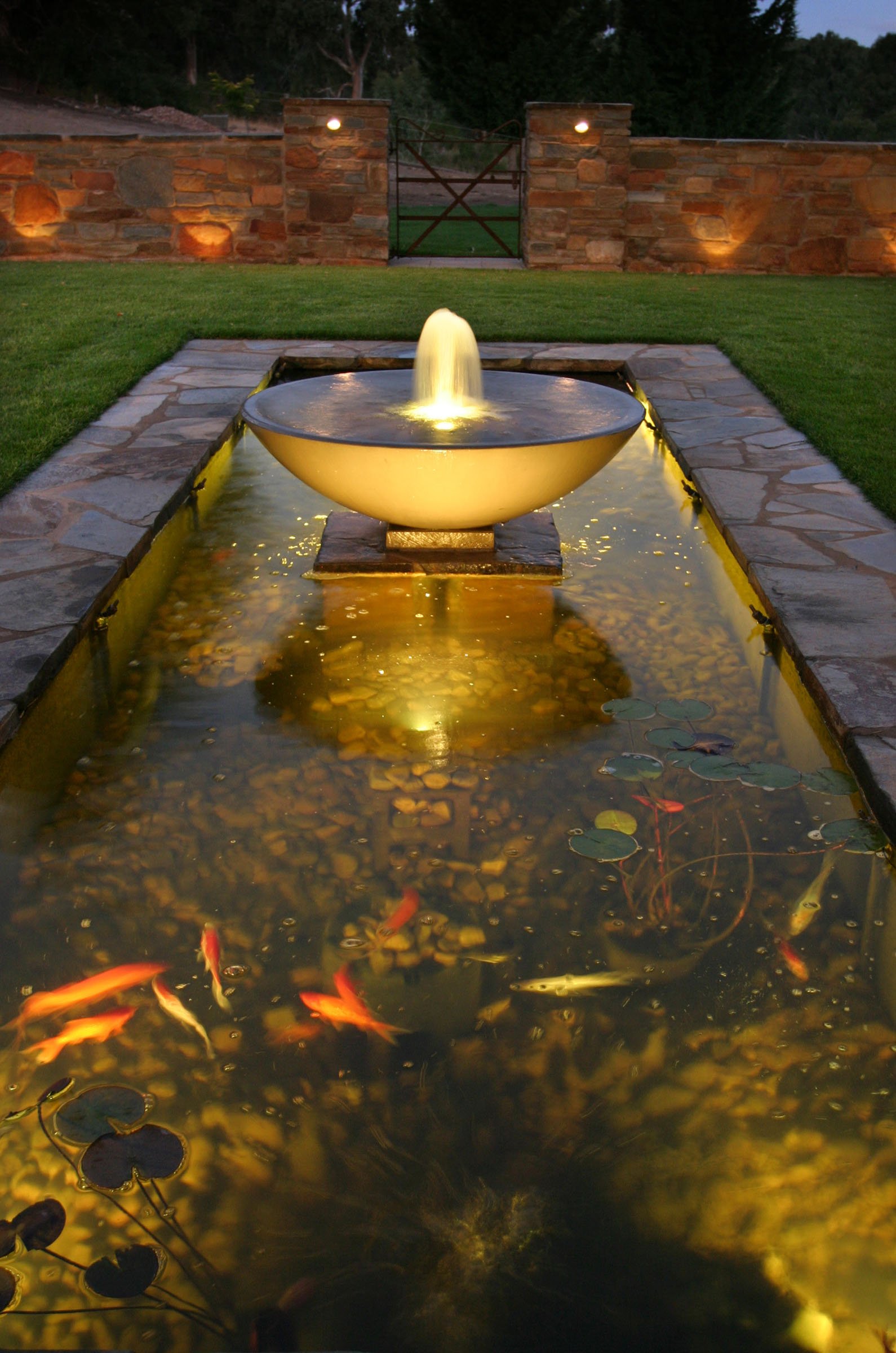 A decorative outdoor water fountain with a bowl-shaped top, illuminated from below, is situated in a narrow rectangular pond filled with koi fish. The pond is bordered by a stone walkway, with a brick wall and a metal gate in the background, illuminated by outdoor lights during dusk.