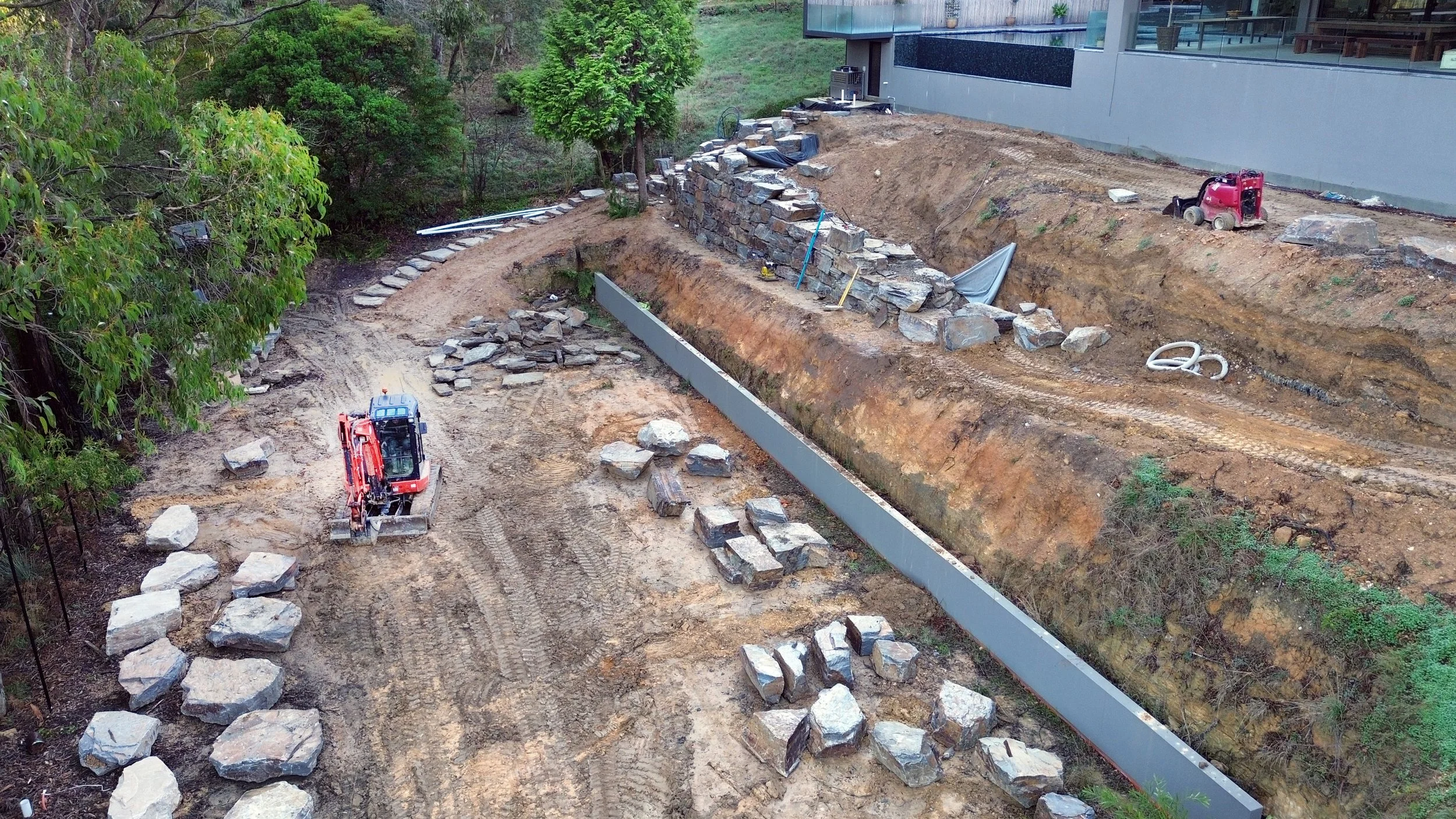 Construction site with excavator and stone wall being built near a modern building, surrounded by trees.