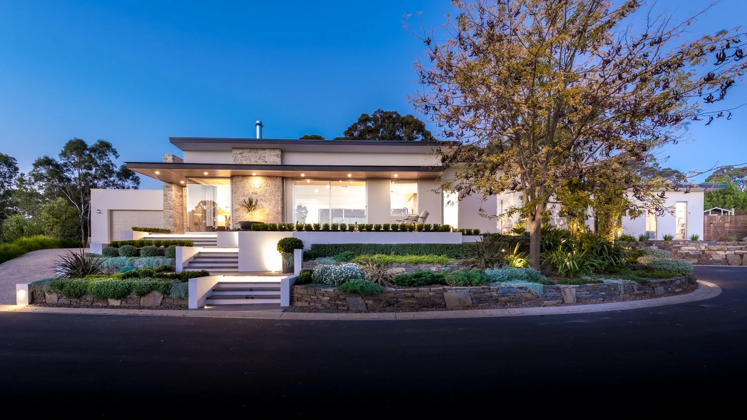 Modern house with large glass windows and stone accents, surrounded by landscaped garden with trees, shrubs, and pathway at dusk.