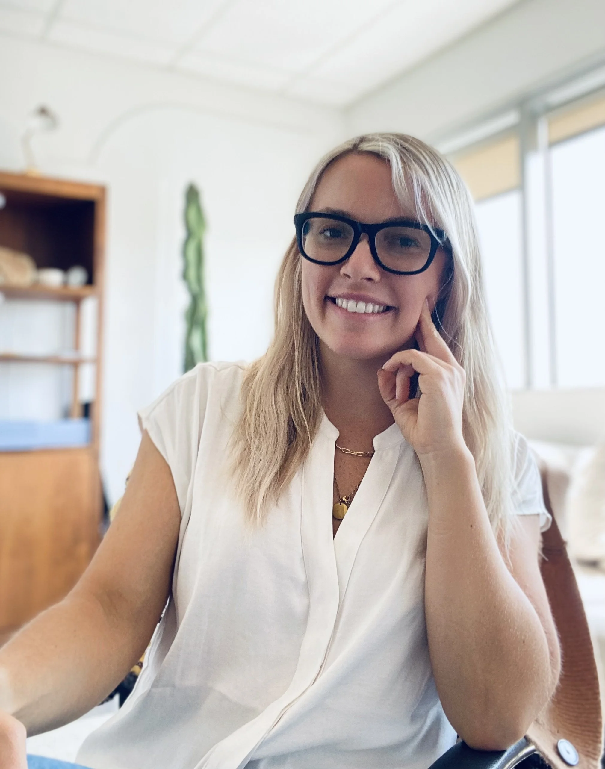 A woman smiling, wearing glasses, sitting indoors near a window with sunlight, wearing a white blouse, with a wooden shelf and a green plant in the background.