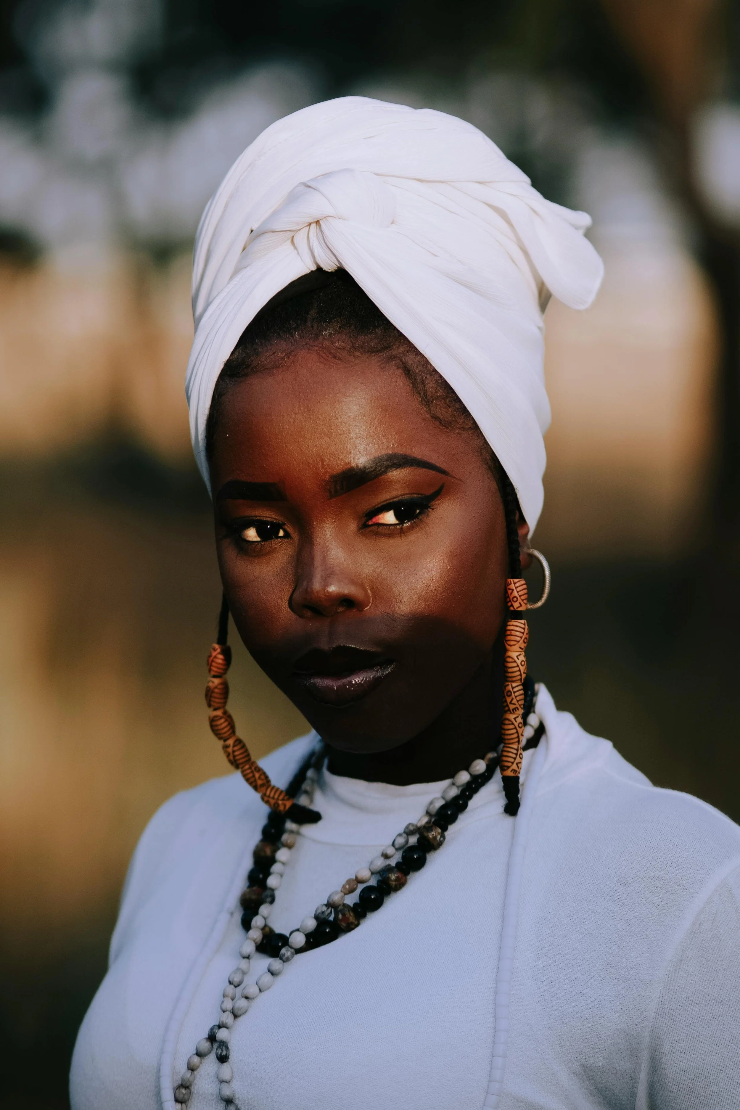A woman with dark skin wearing multiple beaded necklaces and a white headwrap, looking directly into the camera with a neutral expression.