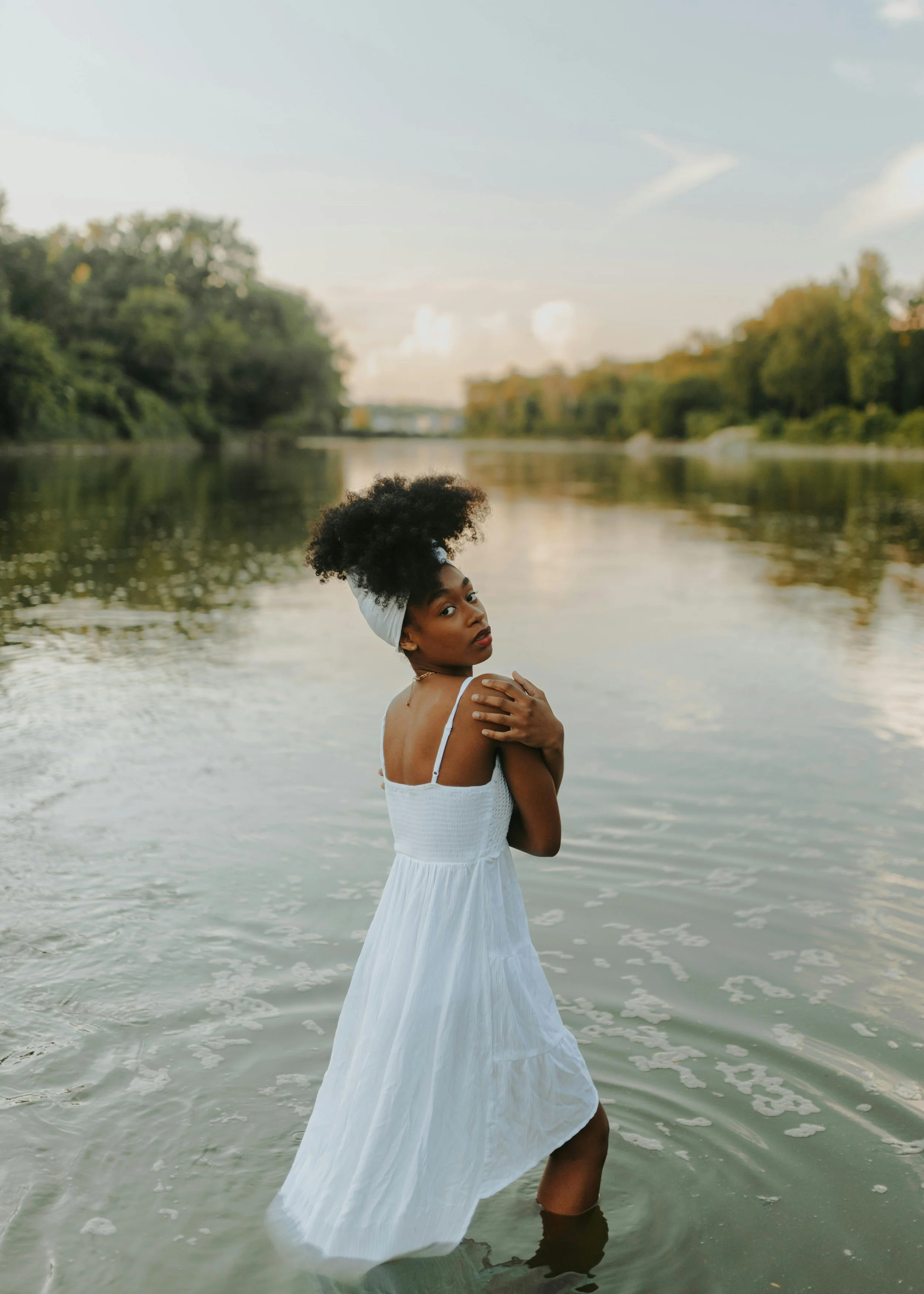 A woman with dark skin and curly hair tied with a white head wrap, wearing a white dress, standing in a shallow river with a background of green trees and a cloudy sky.