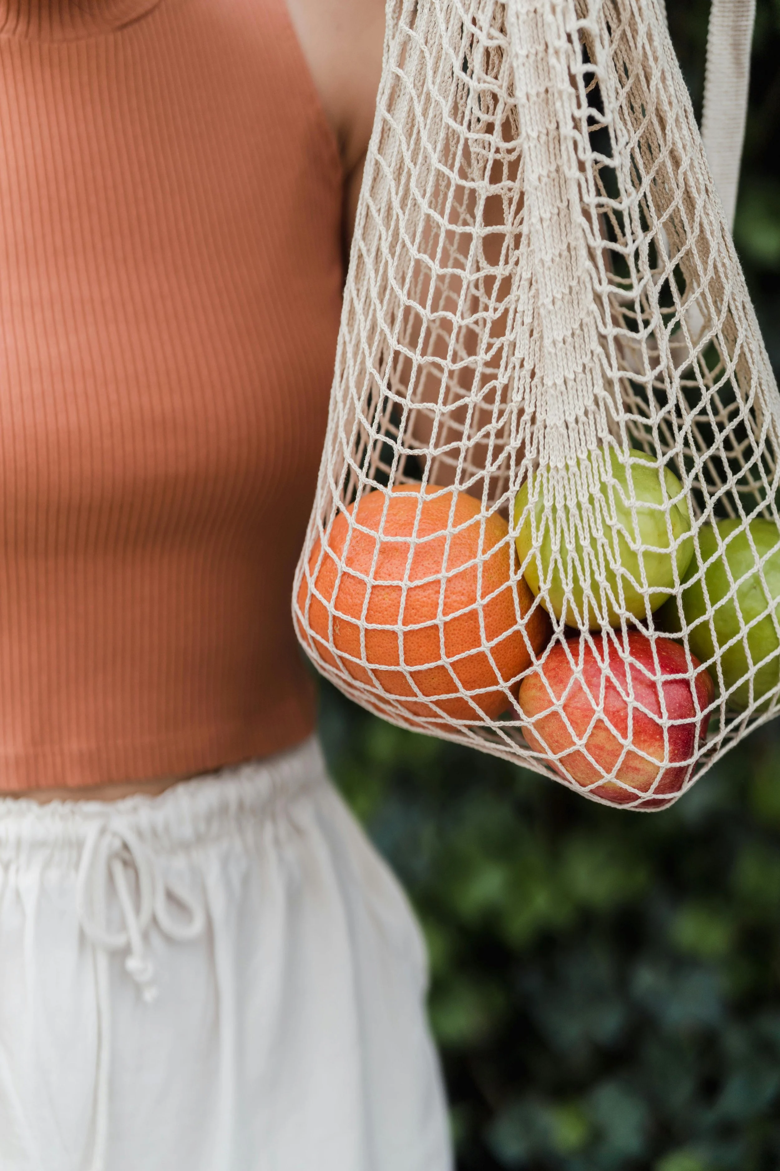 A person wearing a sleeveless orange ribbed shirt and white drawstring skirt holds a white mesh bag with apples inside, set against a leafy background.