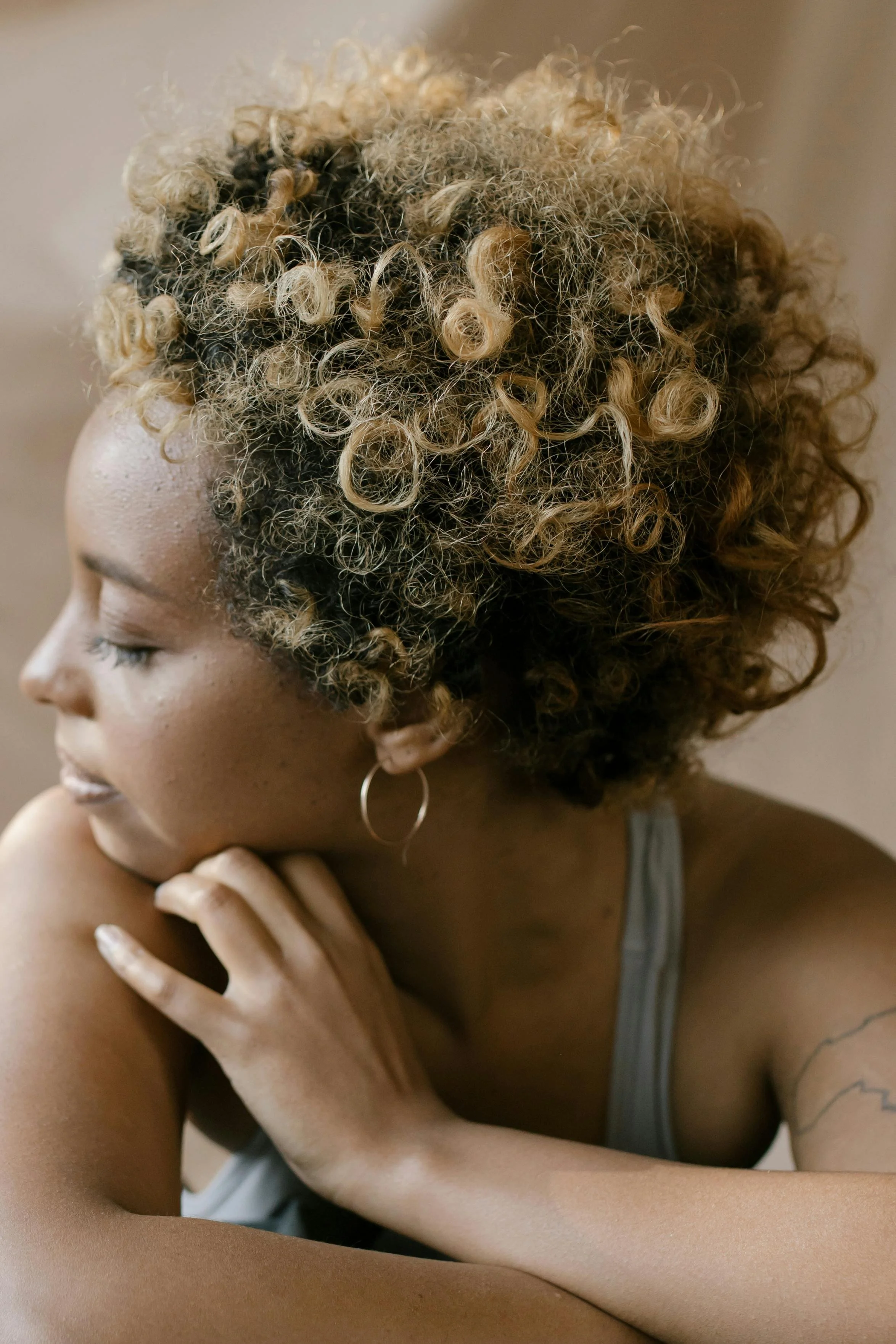A woman with curly blonde hair and a tan skin tone is seen in profile. She has her eyes closed, and one hand gently touches her shoulder. She is wearing a gray tank top and a hoop earring.