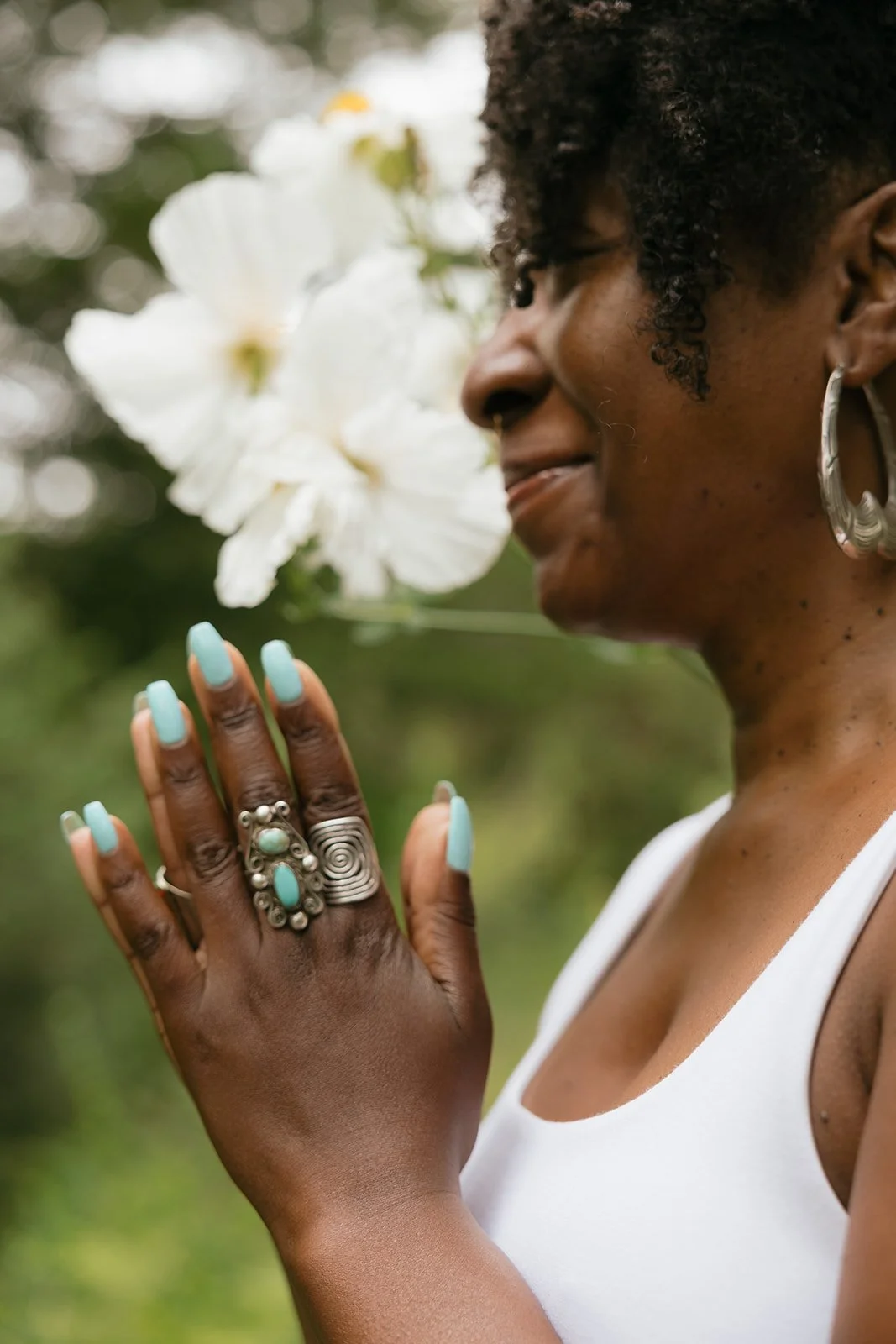 A woman with curly hair and earrings, wearing a white tank top, holding her hand up with rings and turquoise jewelry, smelling white flowers outdoors.