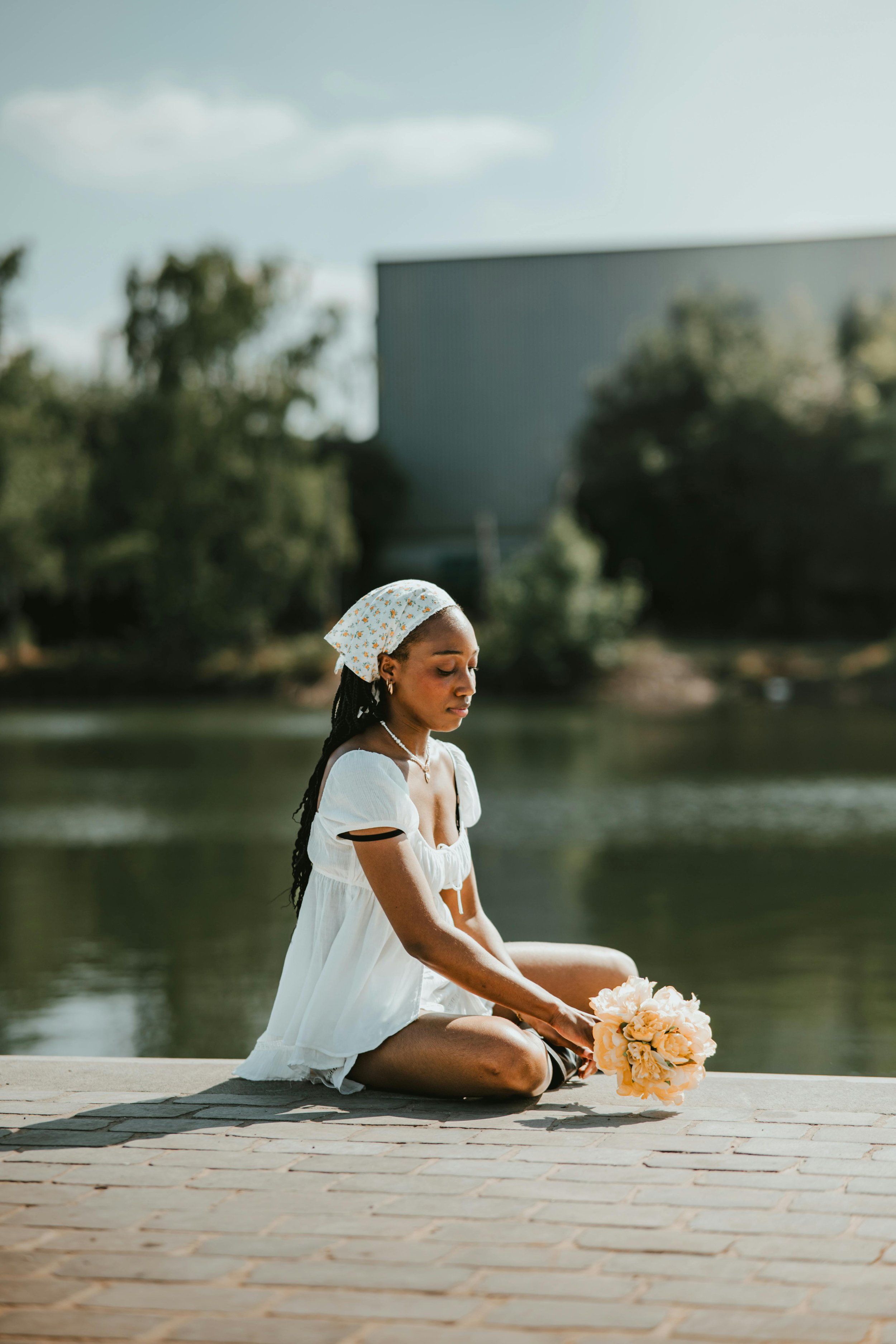 A woman with long dark hair, wearing a white dress and a floral headscarf, sitting cross-legged by a body of water, holding a bouquet of peach-colored flowers.